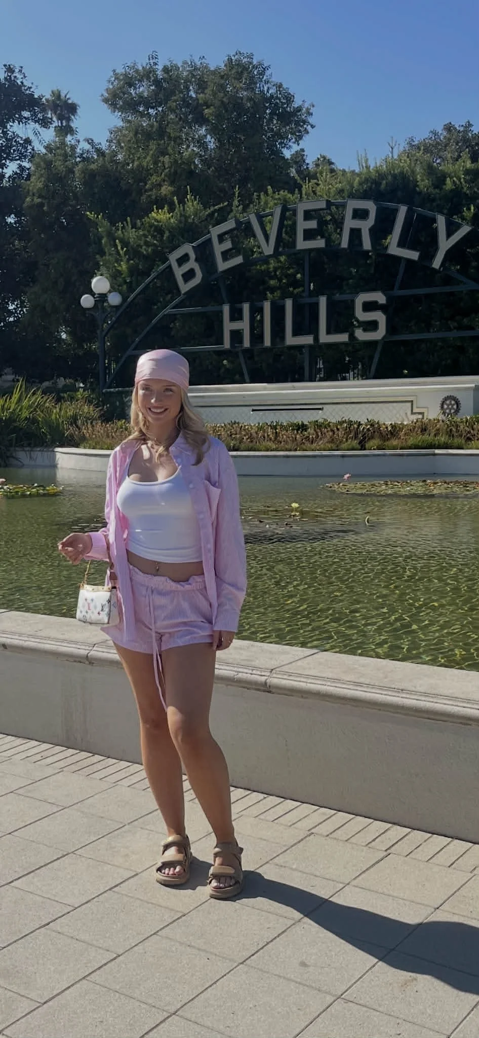 Young woman in pink outfit standing in front of Beverly Hills sign with water feature and trees in background.