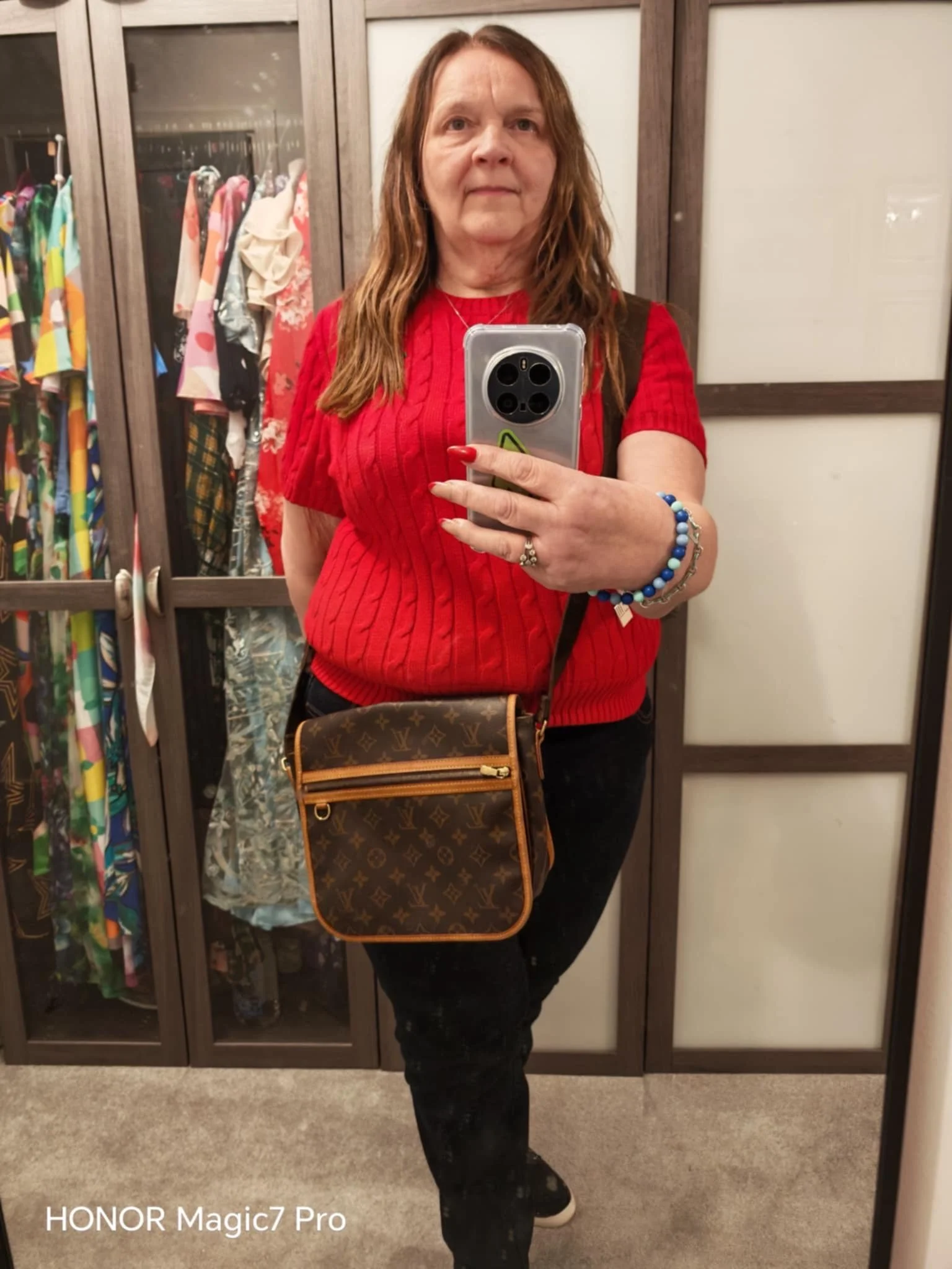 A woman with long, wavy red hair taking a mirror selfie in a fitting room. She is wearing a red short-sleeved sweater, black pants, and has a Louis Vuitton crossbody bag. Behind her are glass-fronted closets with colorful dresses and clothing.