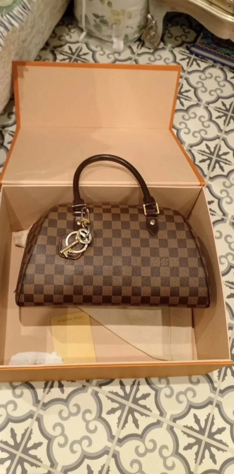 A Louis Vuitton checkered handbag in a brown and beige pattern inside a pink box with an orange border, sitting on a patterned tiled floor.