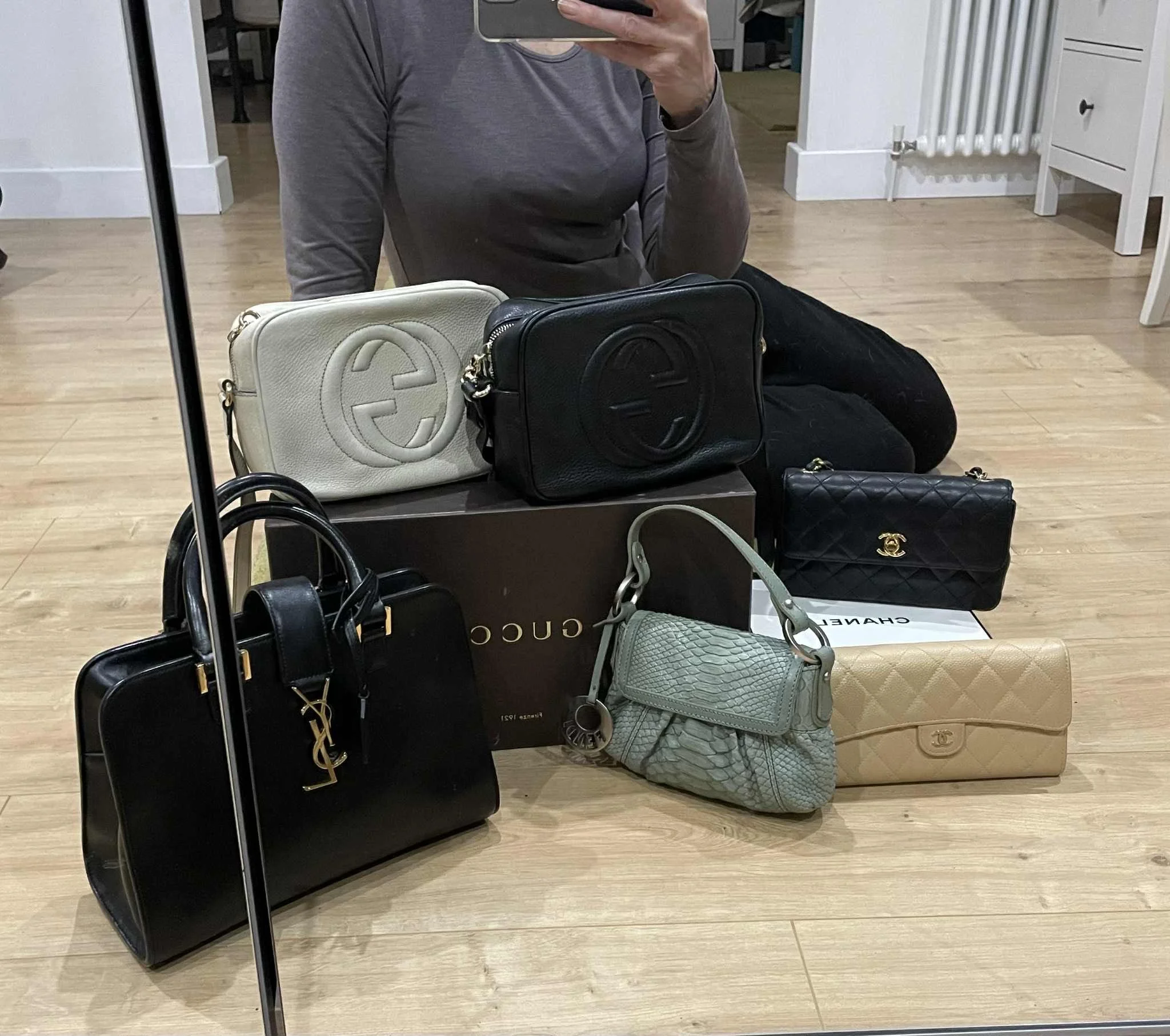 Mirror reflection showing a person taking a photo with several designer handbags on the floor, including Gucci, Chanel, Saint Laurent, and Chanel boxes, in a room with wooden flooring and white furnishings.