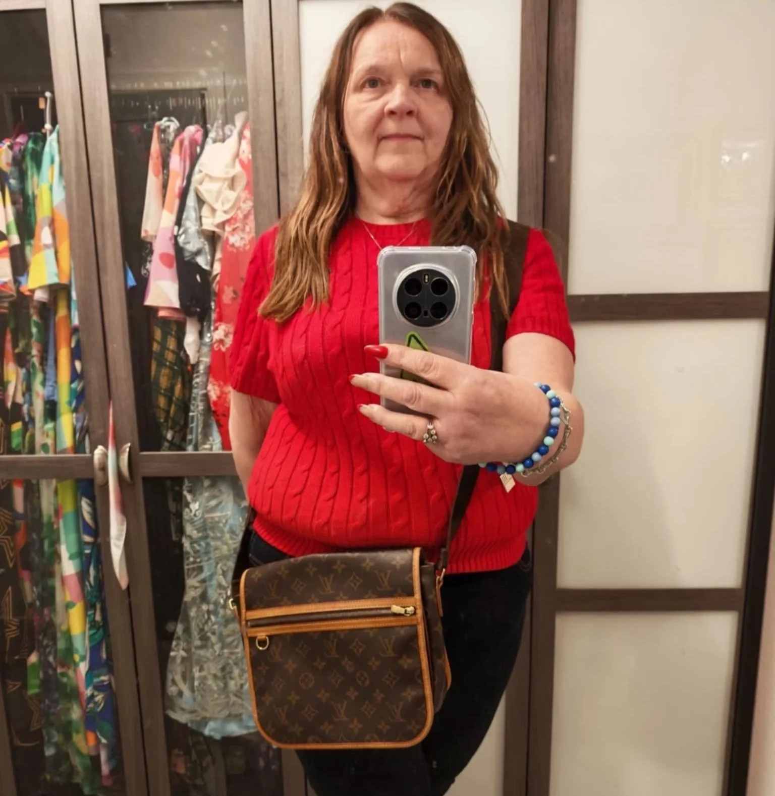 A woman with long red hair wearing a red sweater takes a mirror selfie inside a dressing room. She has multiple bracelets on her right wrist, a Louis Vuitton purse diagonally across her body, and is holding a phone with a silver case. Behind her is a wardrobe with colorful clothes.