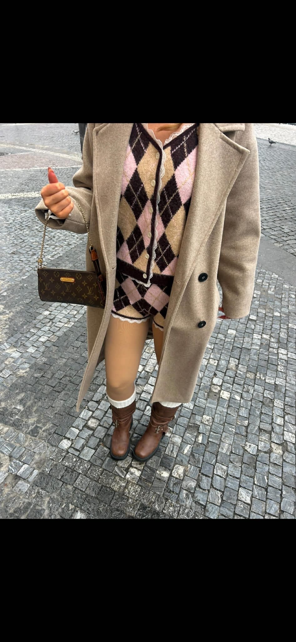 Person wearing a beige coat, pink and black argyle sweater, beige shorts, white socks, and brown boots, holding a Louis Vuitton purse, standing on a cobblestone street.