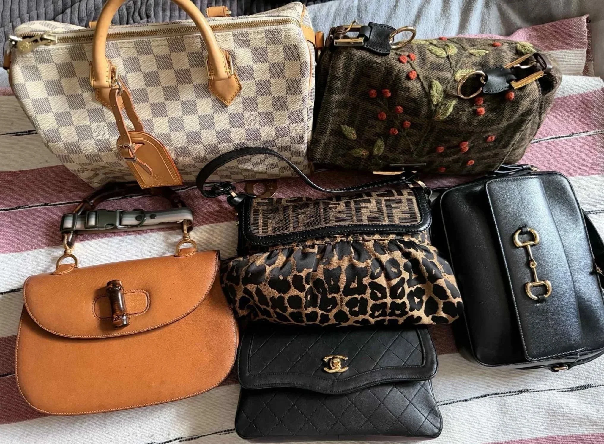 Collection of seven handbags placed on a striped cloth surface, including designs from Louis Vuitton, Fendi, Gucci, and Chanel, featuring various patterns, colors, and styles.