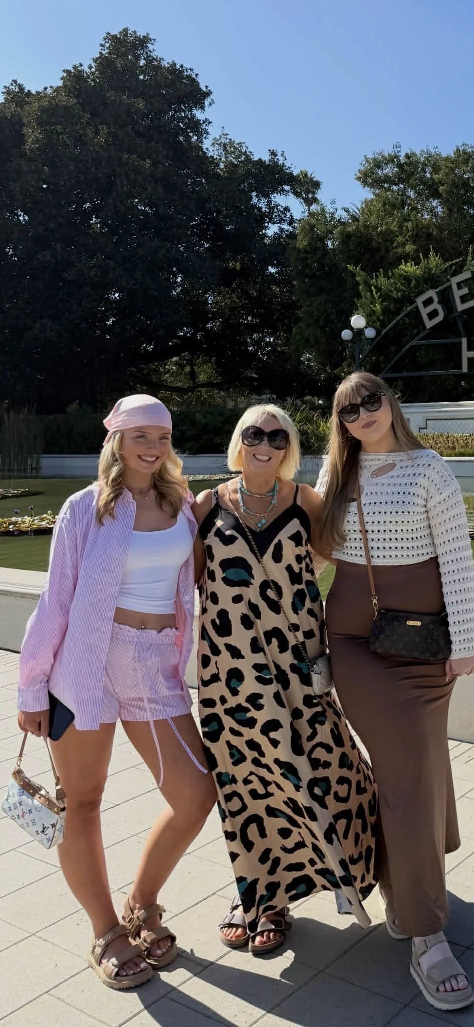 Three women standing outdoors in front of a pond, smiling, with large trees behind them. The woman on the left is wearing a pink hat, a striped pink shirt, matching pink shorts, and sandals. The woman in the middle is wearing a leopard print dress with black, beige, and blue patterns, with sunglasses and multiple necklaces. The woman on the right is wearing a white cropped sweater, brown skirt, sunglasses, and carrying a small black purse.