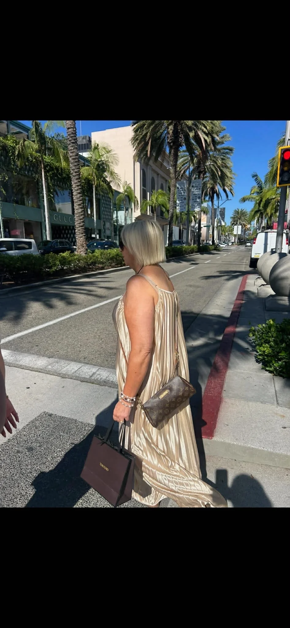 A woman with blonde hair in a shoulder-length bob, wearing a shiny beige dress, walking on a sunny street with palm trees and luxury storefronts in the background, holding shopping bags and a Louis Vuitton purse.