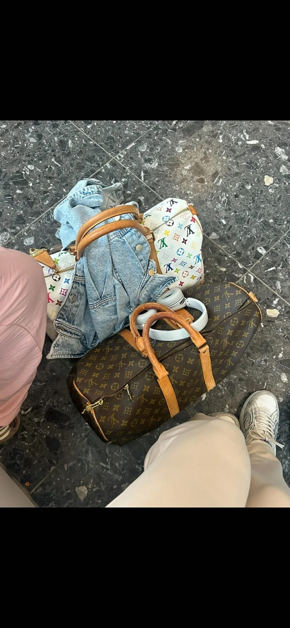 Luggage and personal belongings on the floor, including a Louis Vuitton duffel bag, a multicolored Louis Vuitton bag, a denim jacket, a light pink garment, a pair of white headphones, and a person's legs and shoes.