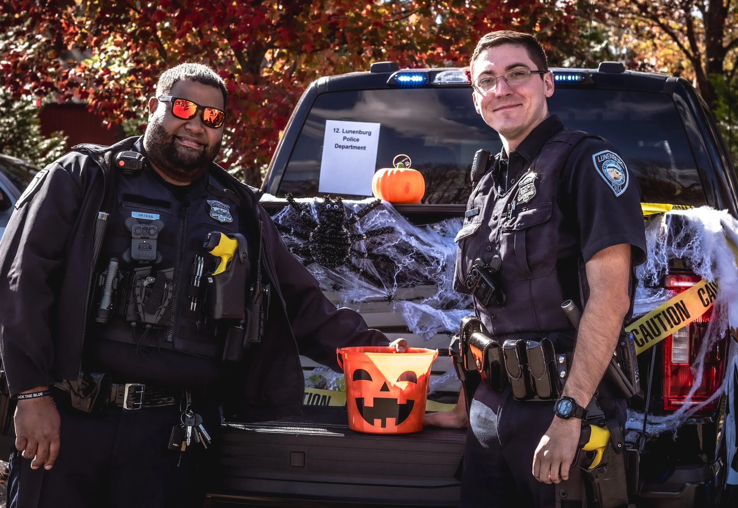 Two police officers standing in front of a police vehicle decorated with Halloween decorations, including a pumpkin, spider web, and caution tape.