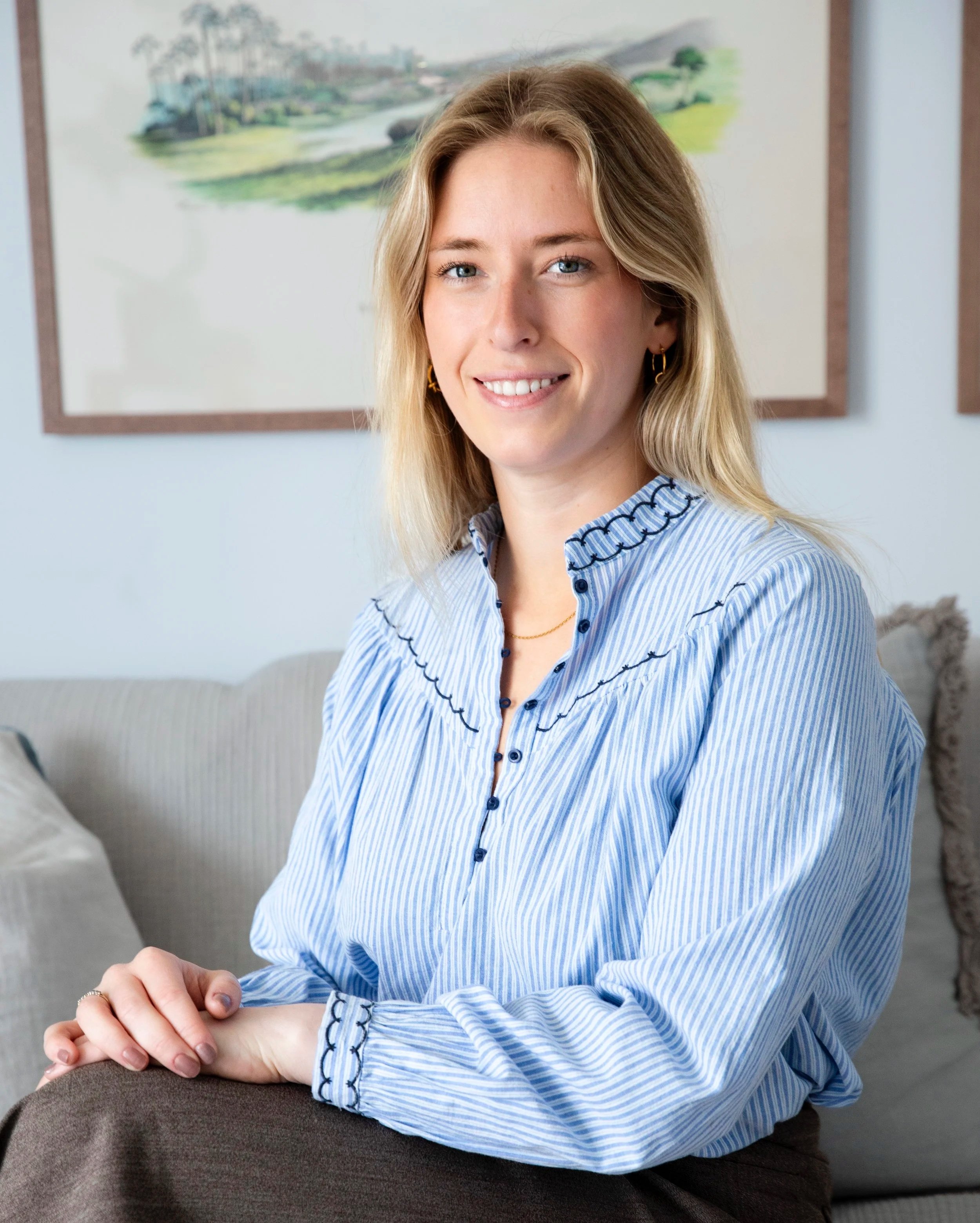 A young woman with blonde hair, blue eyes, and hoop earrings, smiling and sitting with her arms on a table. She wears a blue and white striped blouse. Behind her is a framed landscape painting and a light-colored wall.