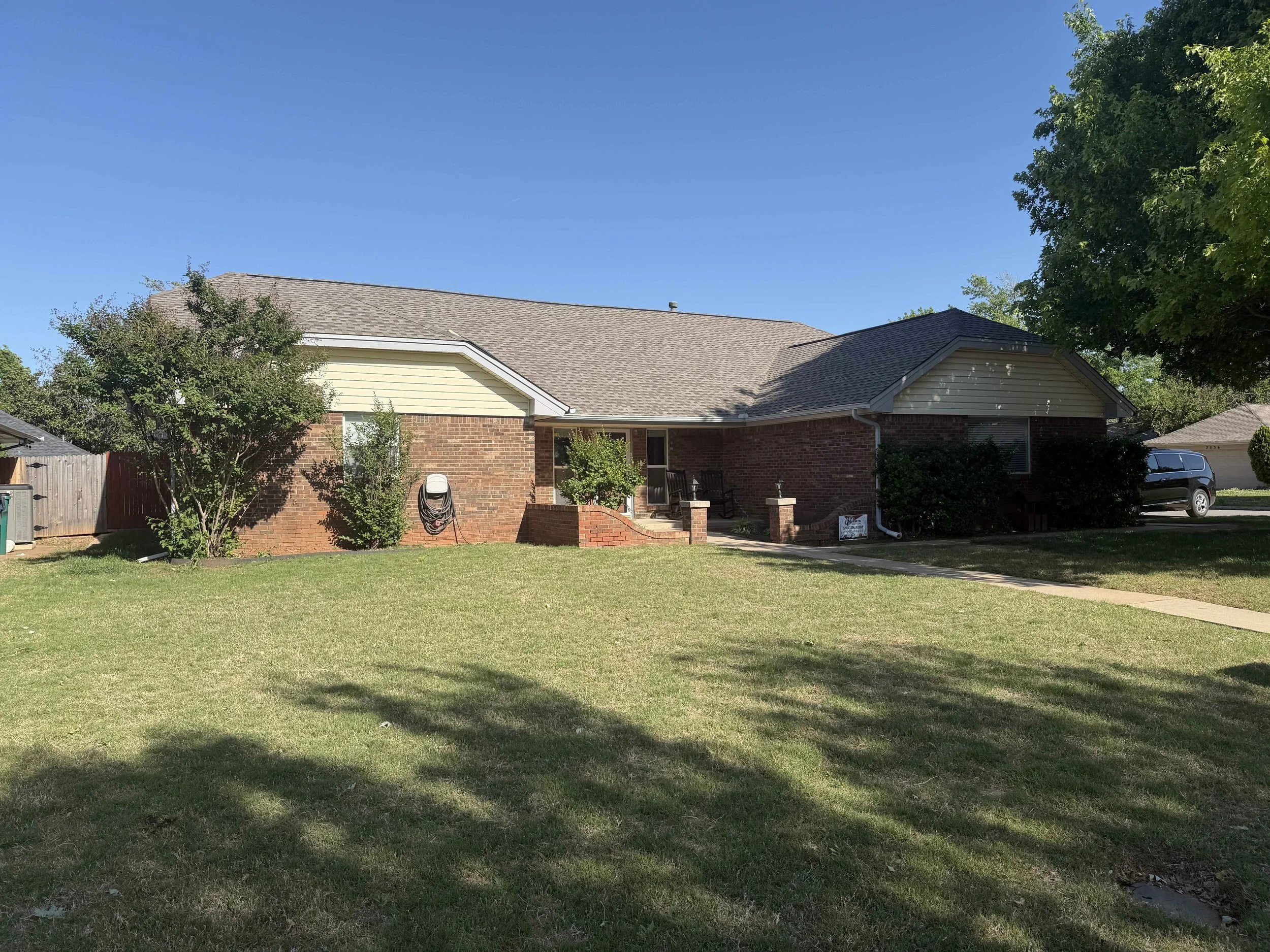 A suburban house with brick and siding exterior, a new sloped roof, front porch with stairs, green lawn, trees, a car parked on the driveway, and a clear blue sky.