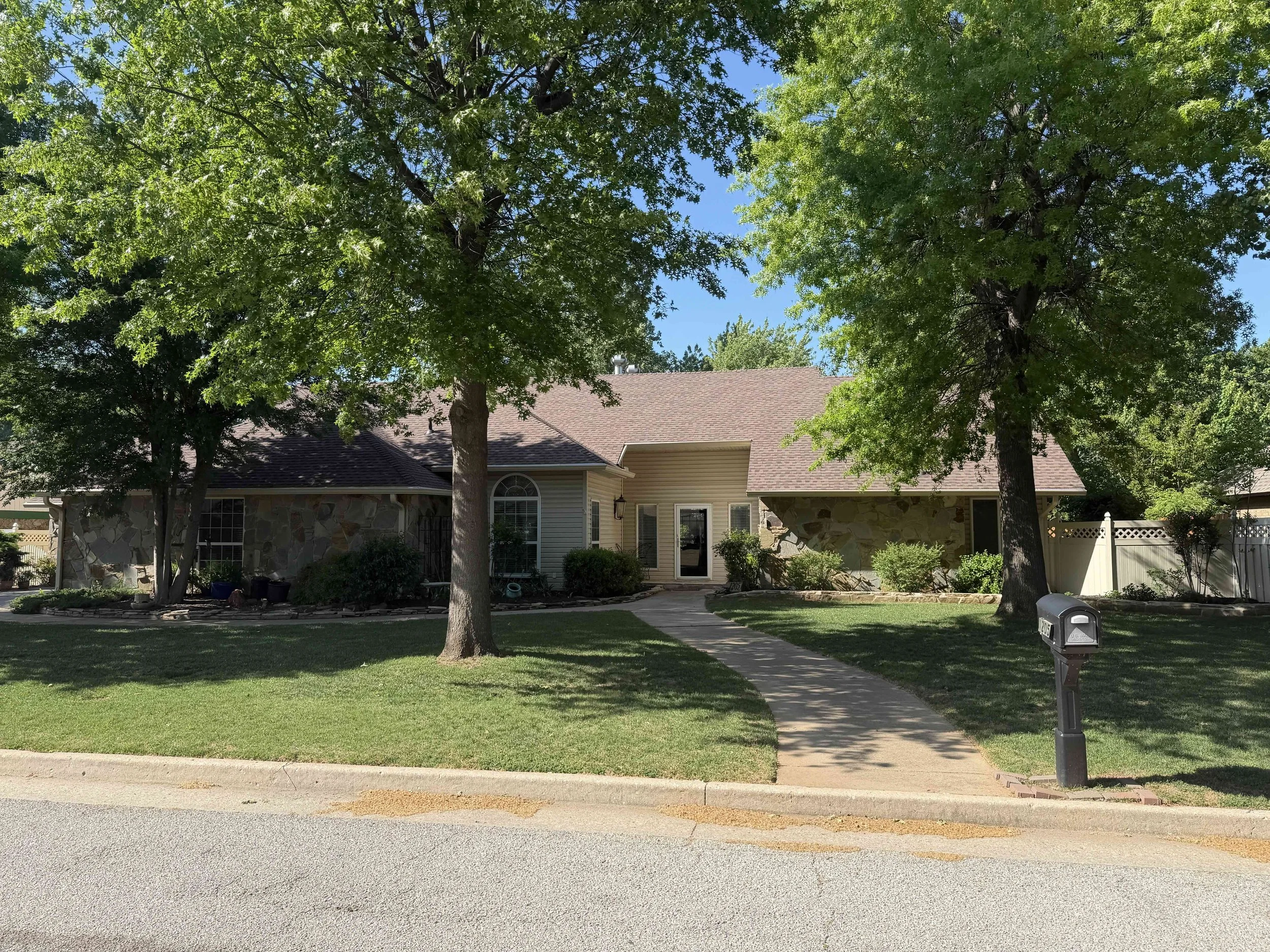 A house with a brick and stone exterior, large windows, and a new brown roof, surrounded by green trees and a well maintained lawn, with a sidewalk and a black mailbox in the front yard.