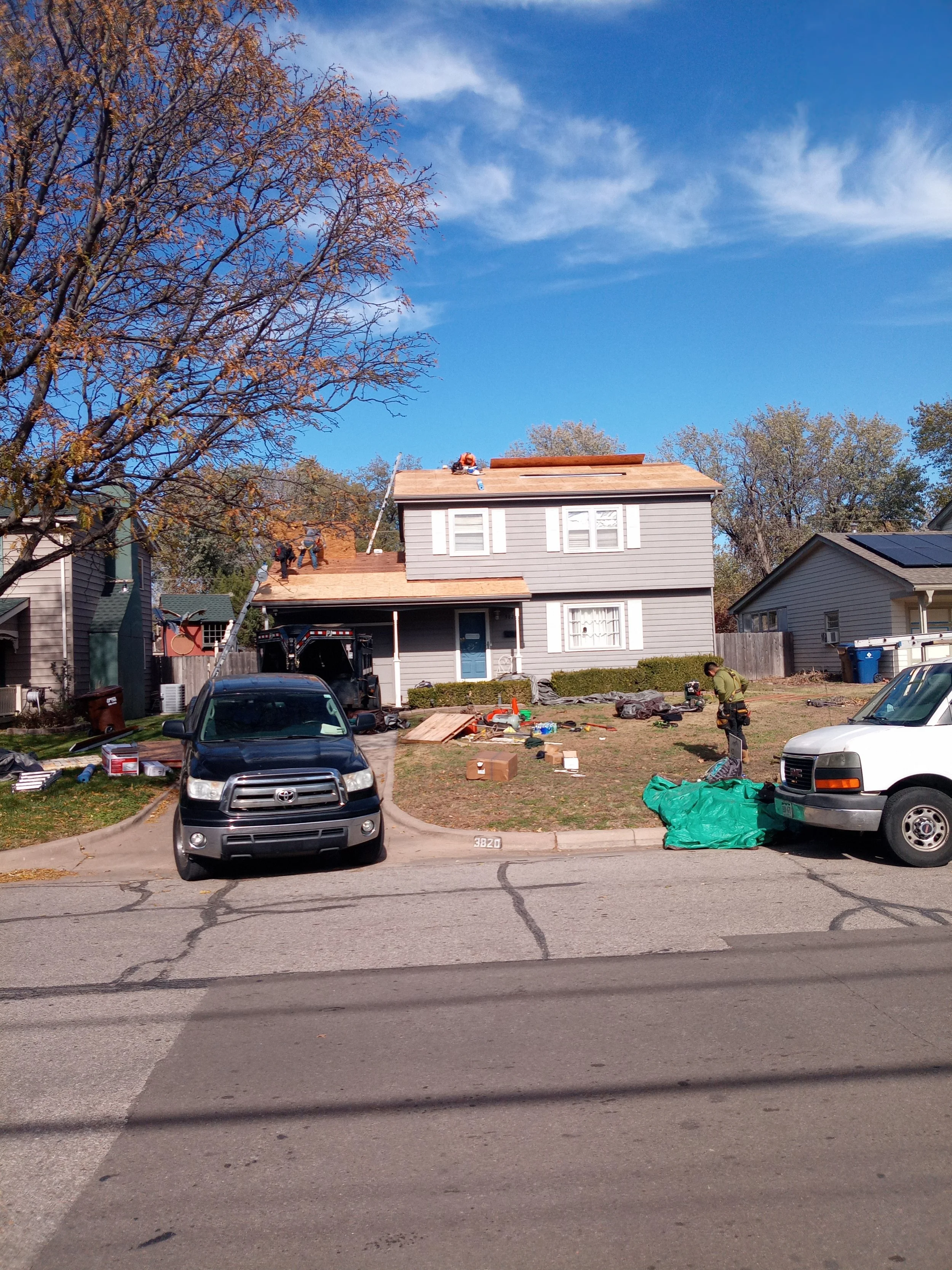 Two men working on the roof of a gray twostory house with white shutters under a blue sky with scattered clouds. The house has a wooden boarded section on the roof, and construction tools and materials are scattered in the front yard. Two vehicles ar