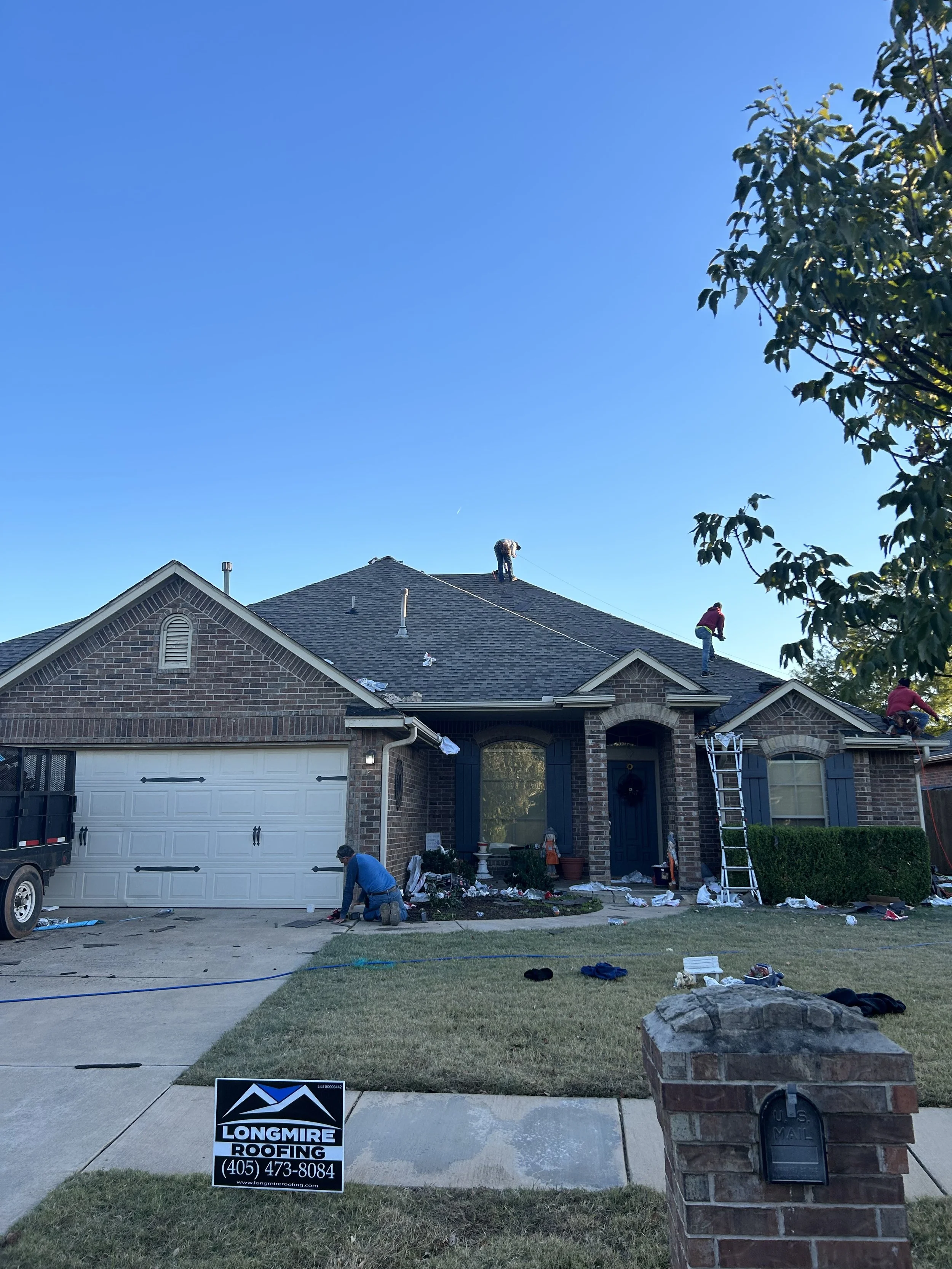 House with workers repairing or replacing the roof, tools and materials scattered on the driveway and lawn, a sign for Longmire Roofing in front of the house, and a brick mailbox in the foreground.