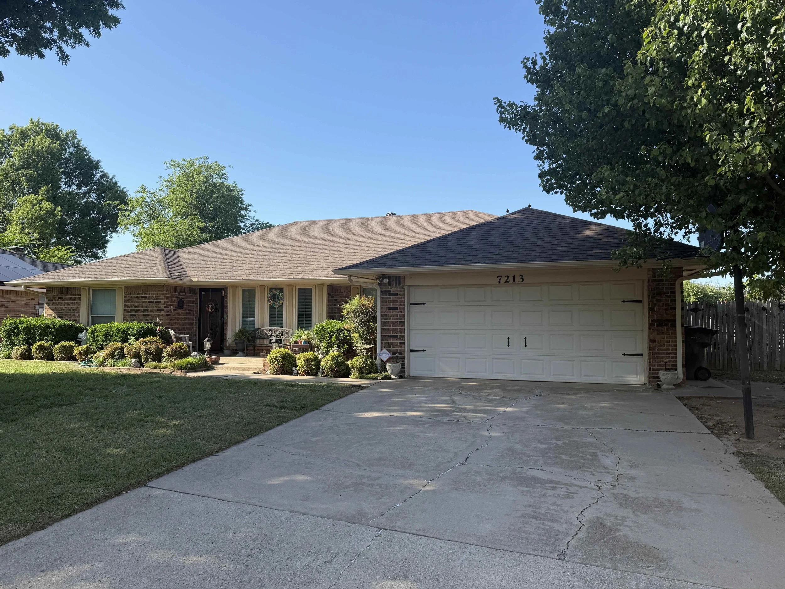 Front view of a single story brick house with new gray shingle roof, white garage door, and a small porch with patio furniture. The front yard has green grass, shrubs, and trees on both sides. A concrete driveway leads to the garage.