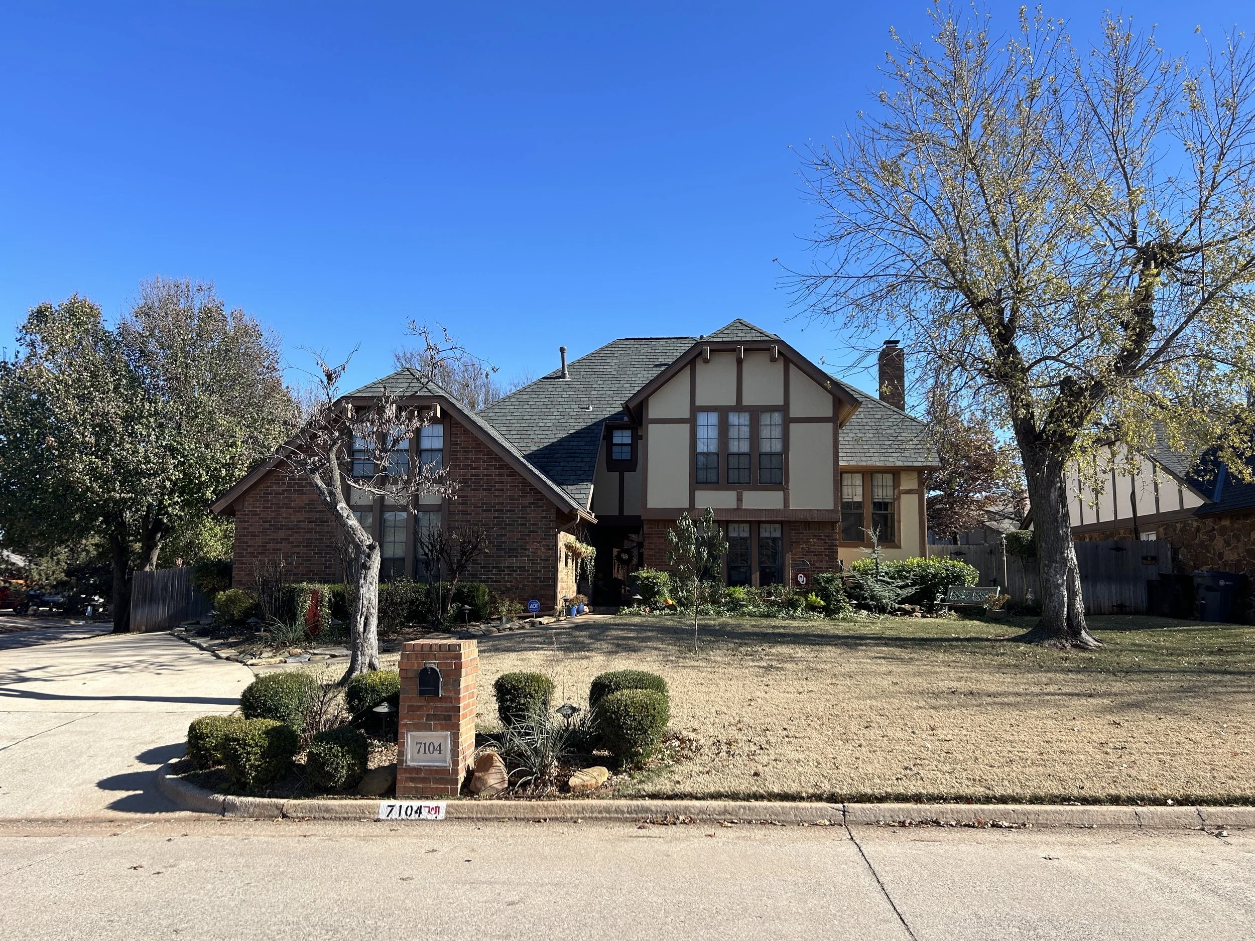 Front yard of a house with a brick and stucco exterior, multiple trees, a mailbox, and a driveway under a clear blue sky.