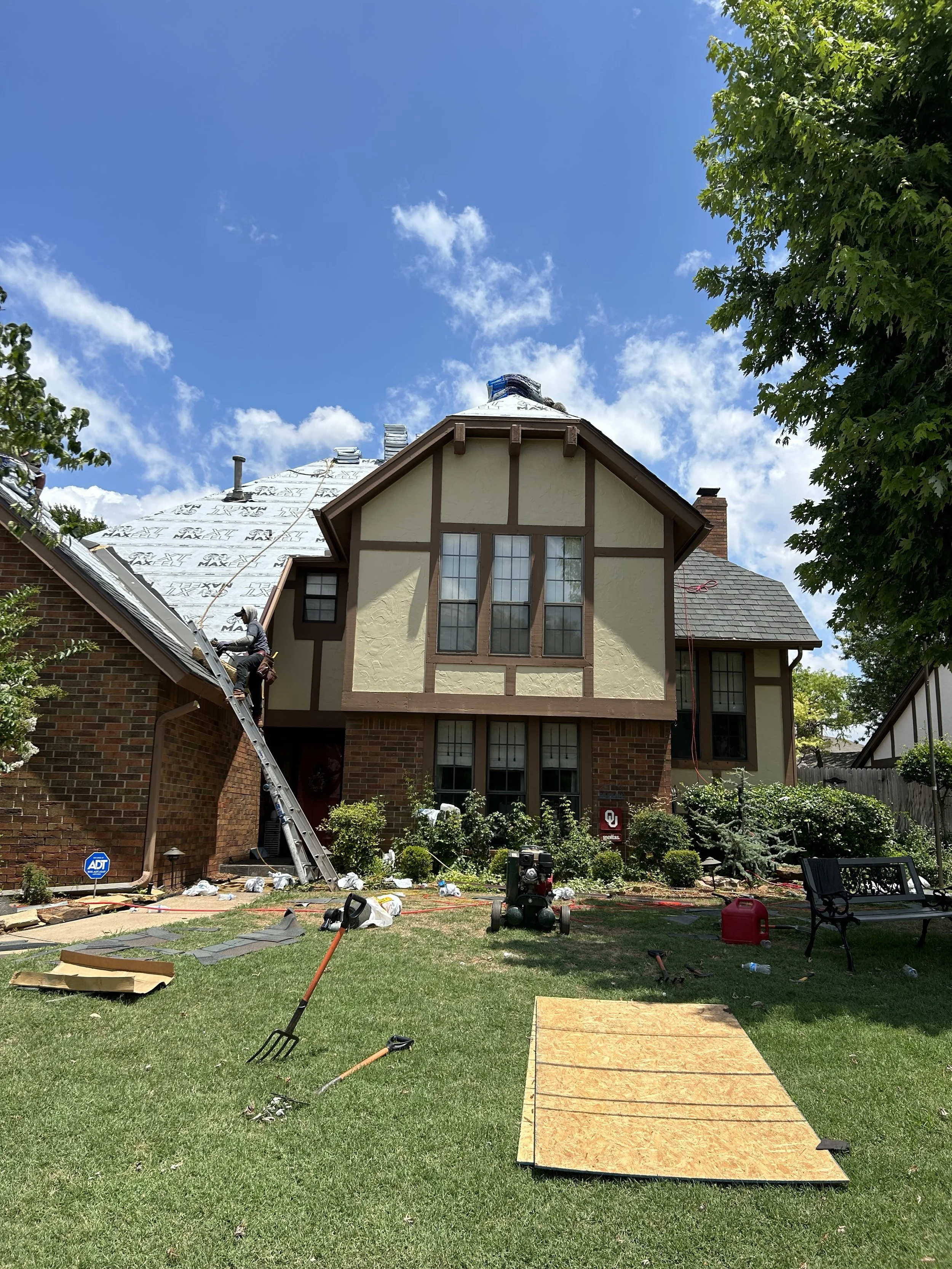 Backyard of a house under construction with workers working on the new roof, a ladder, tools, and construction materials scattered on the lawn. The house has a Tudor-style design with exposed wooden beams and a covered porch.