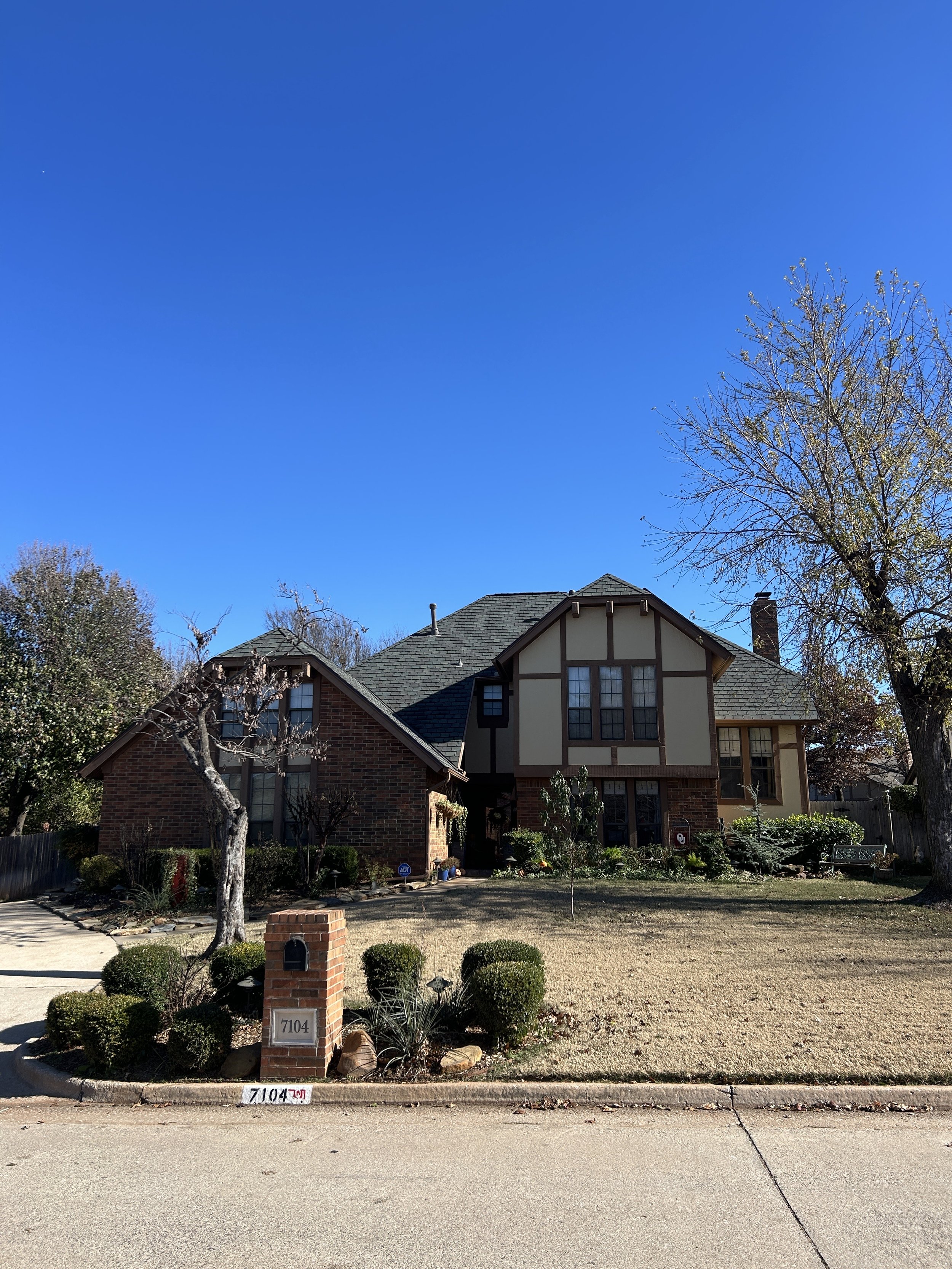 A two story house with a new roof, brick and tan exterior, surrounded by a front yard with bushes, trees, and a brick mailbox, under a clear blue sky.