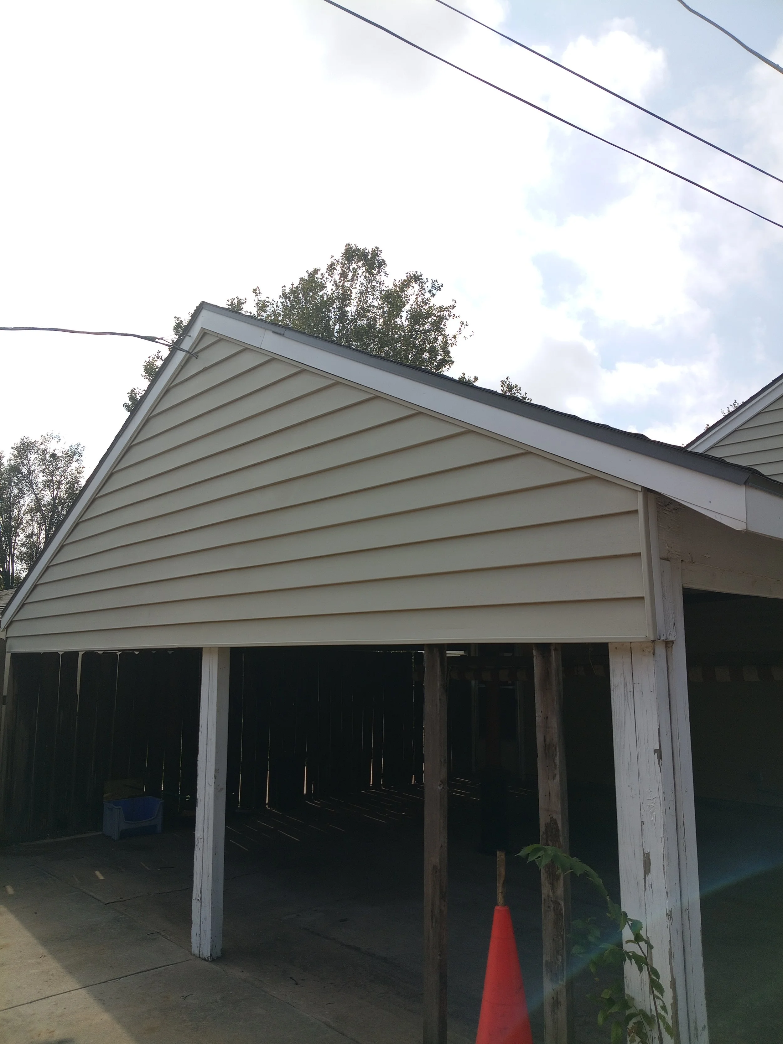 A house with beige siding and a new gabled roof, under a partly cloudy sky, with some electrical wires overhead and a small orange cone near a vine plant at the front.