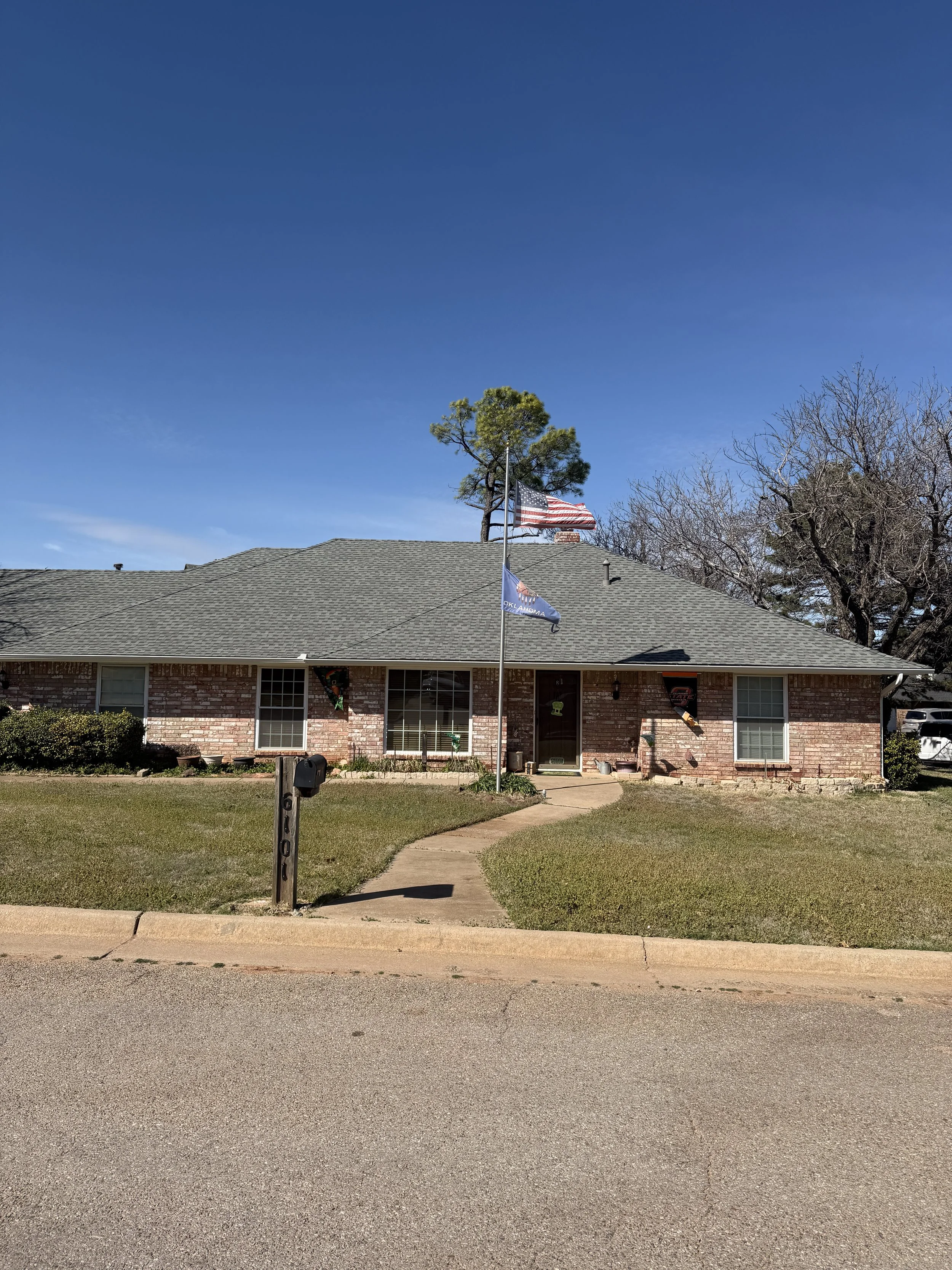 A single-story brick house with a gray shingle roof and a small front yard. The yard has a narrow concrete walkway leading to the front door. There are two windows on the house, one to the left and one to the right of the front door, with white window frames. A black mailbox is mounted on a wooden post near the sidewalk. The house has a flagpole with an American flag and an Oklahoma state flag flying in the yard. There are trees behind the house, with the one on the left being green and the one on the right leafless. The sky above is clear and blue.