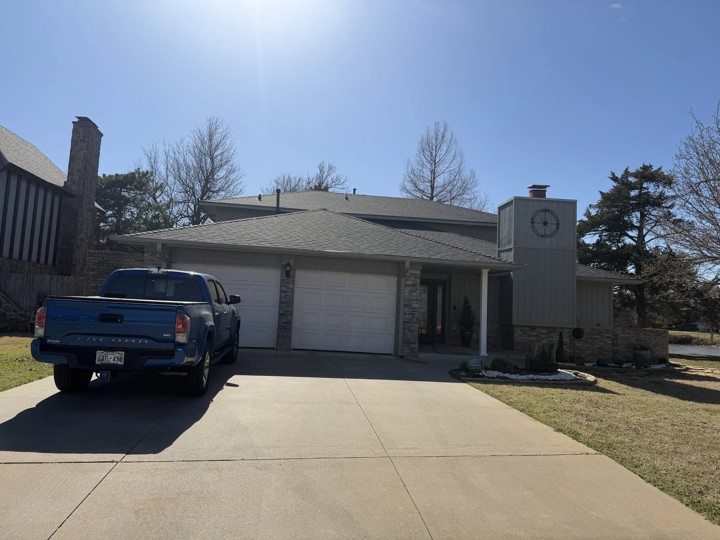 A residential house with a driveway, parked blue pickup truck, and a garage. The house has a new roof, a chimney, a clock on the exterior wall, and trees in the background.