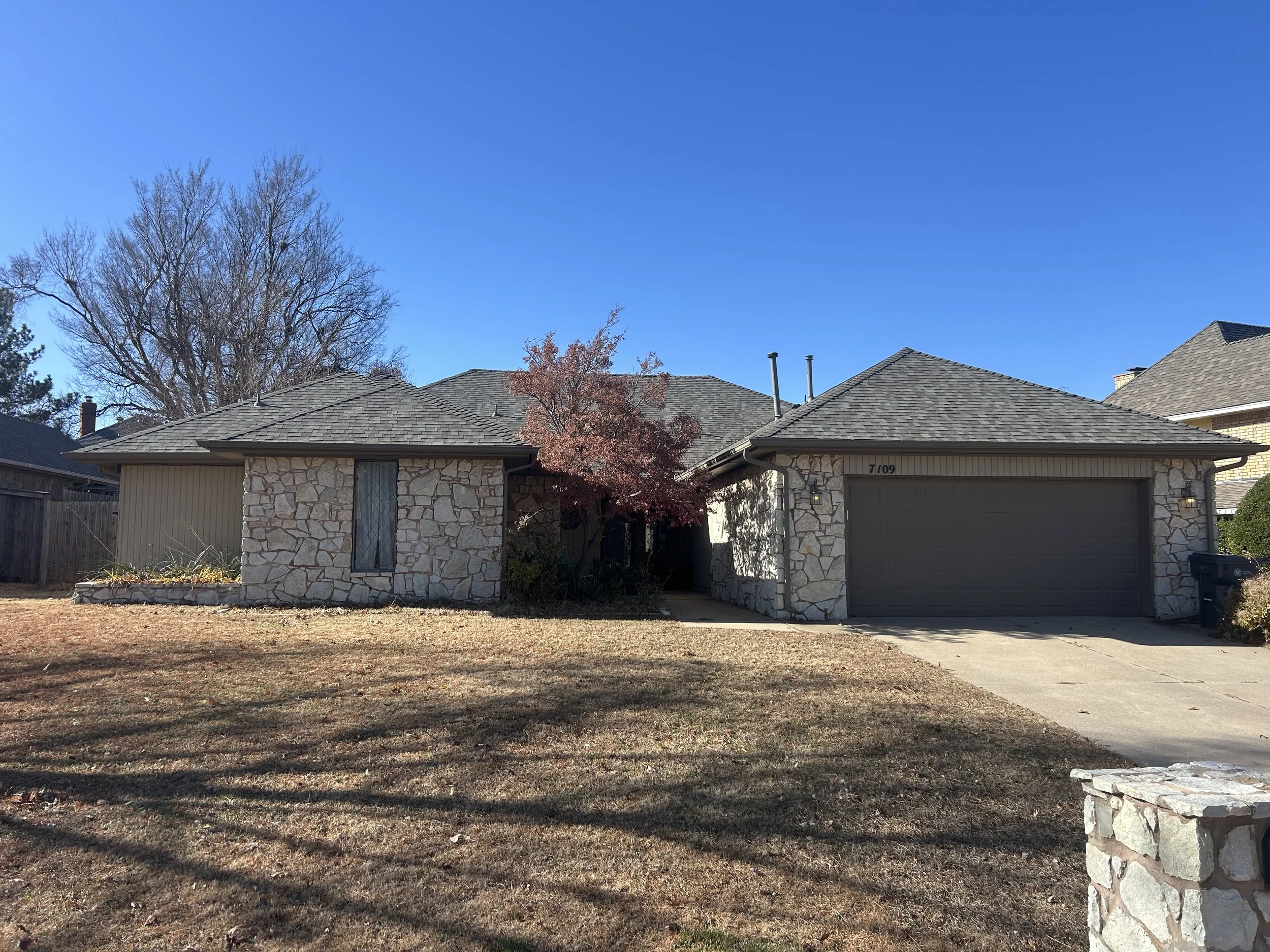 Front view of a house with a new roof, stone and siding facade, a garage door, and a front yard with patchy grass and trees, under a clear blue sky.