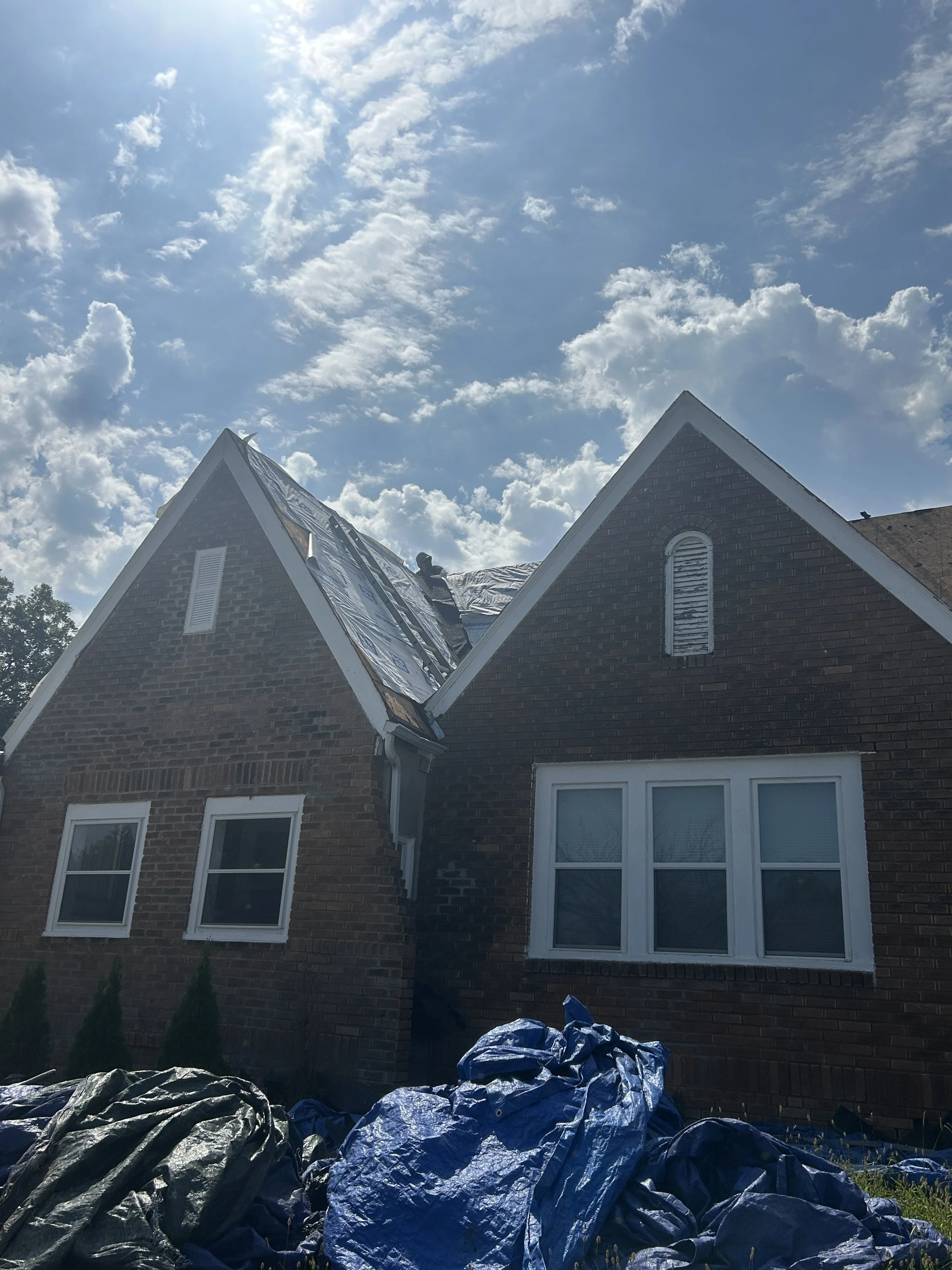 A house with two new gable roofs under a partly cloudy sky, with a worker on the roof installing or repairing solar panels, and black and blue tarps or covers on the ground in front.