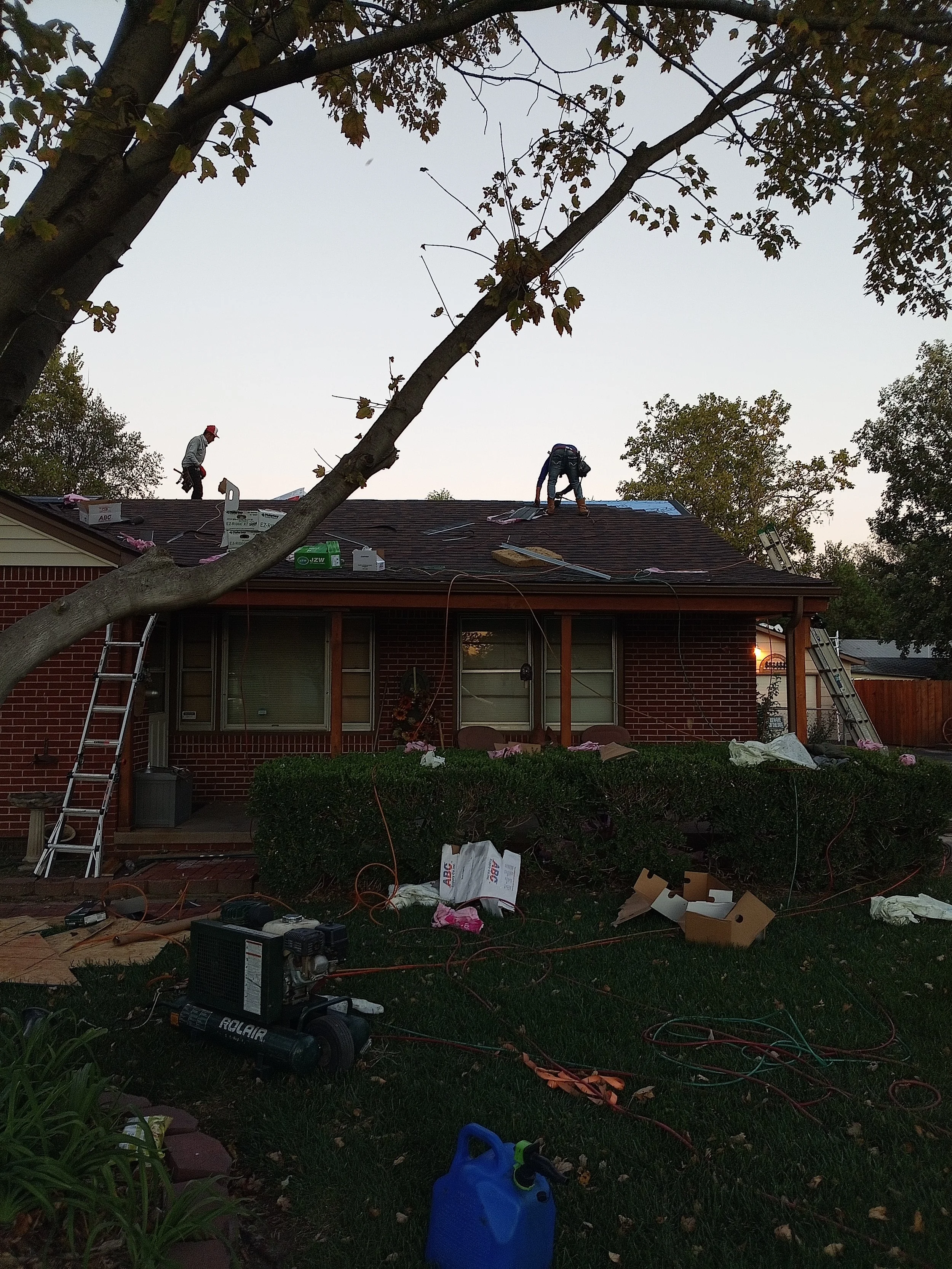 Workers installing a new roof on a house during the evening, with tools, ladder, and roof materials scattered around, and a large tree partially obscuring the view.