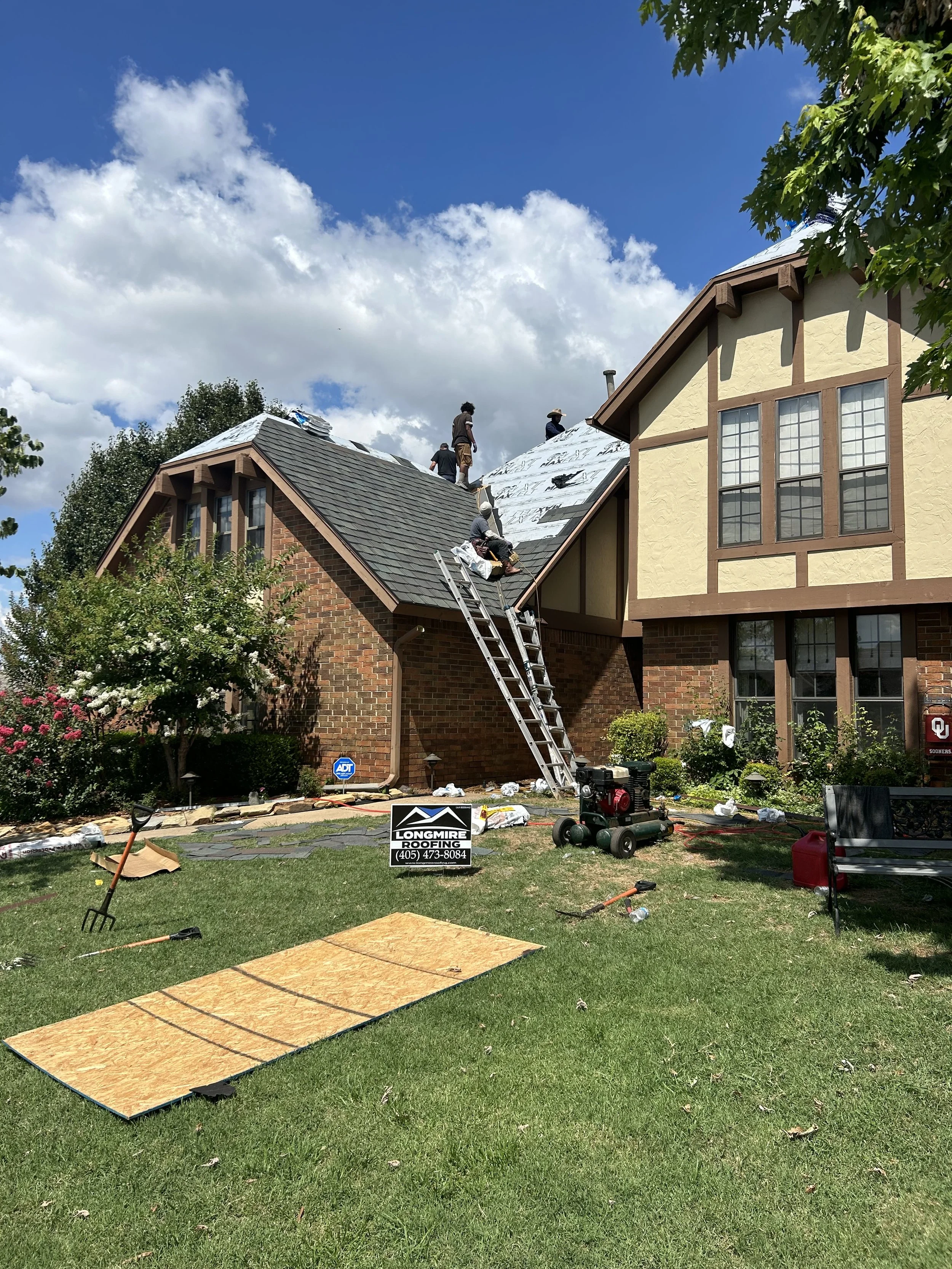 Workers installing a new roof on a house, with tools and materials on the lawn and a sign for Longmire Roofing in the yard.