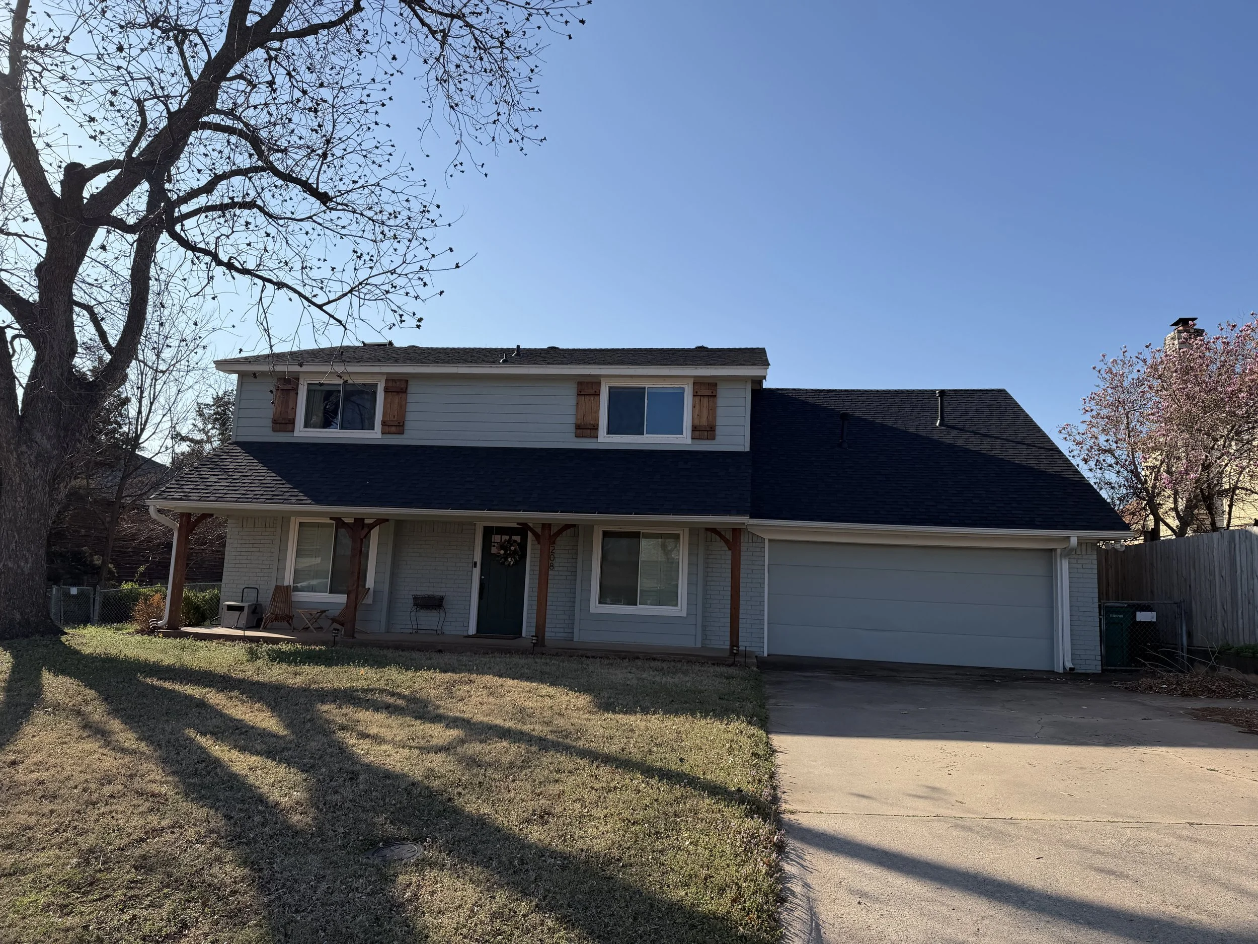 The photo shows the outside of a two story house with light siding, a new dark shingle roof, and a small porch. There are a few trees with no leaves and a flowering tree with pink blossoms. The sky is clear and blue.