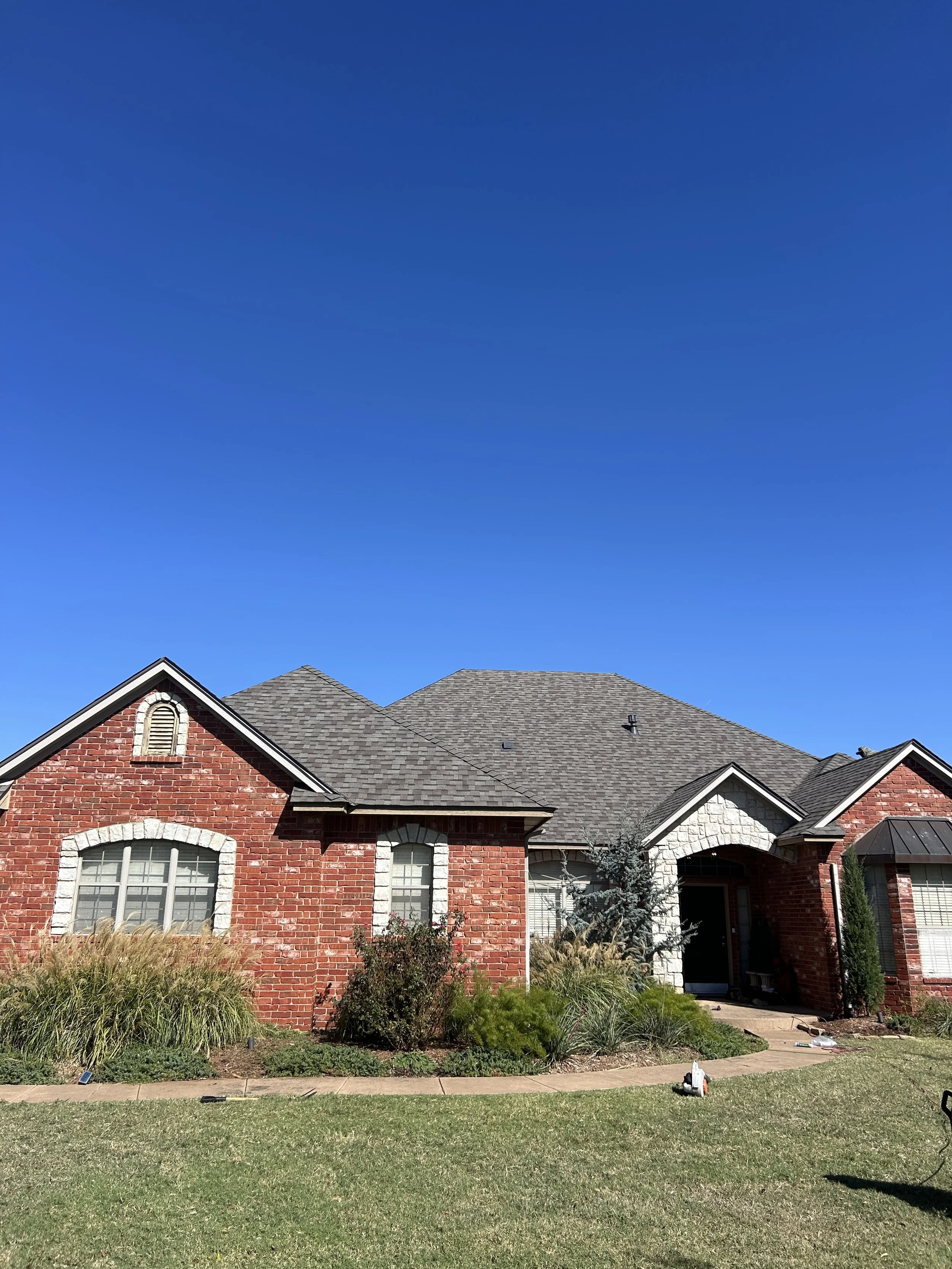 Front view of a brick house with a new gray shingled roof, white window trims, landscaping, and a clear blue sky