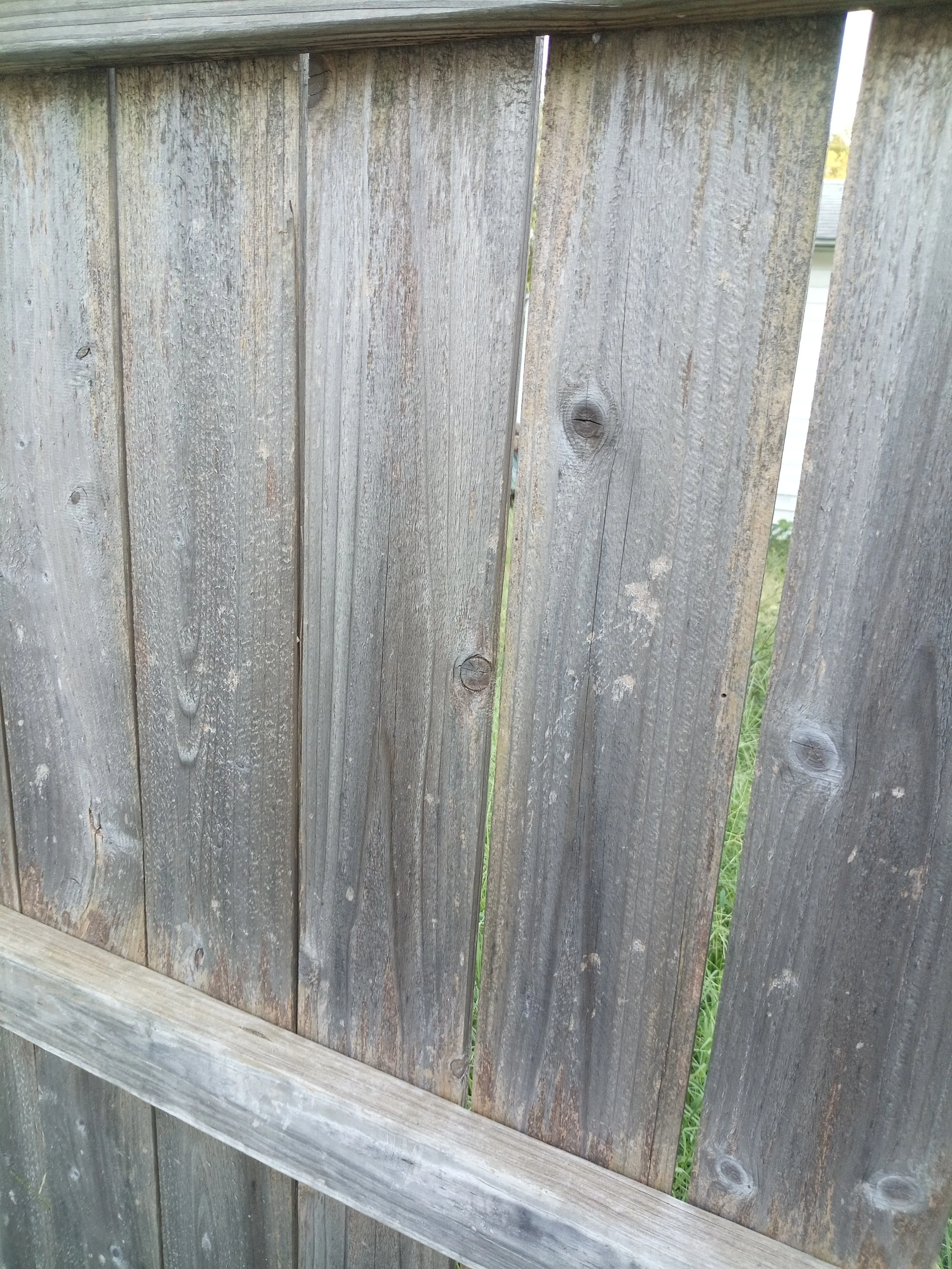 A close-up of a weathered wooden fence needing fence replacement in OKC metro, with vertical planks and visible knots, showing a narrow gap between two planks, with grass visible through the gap.