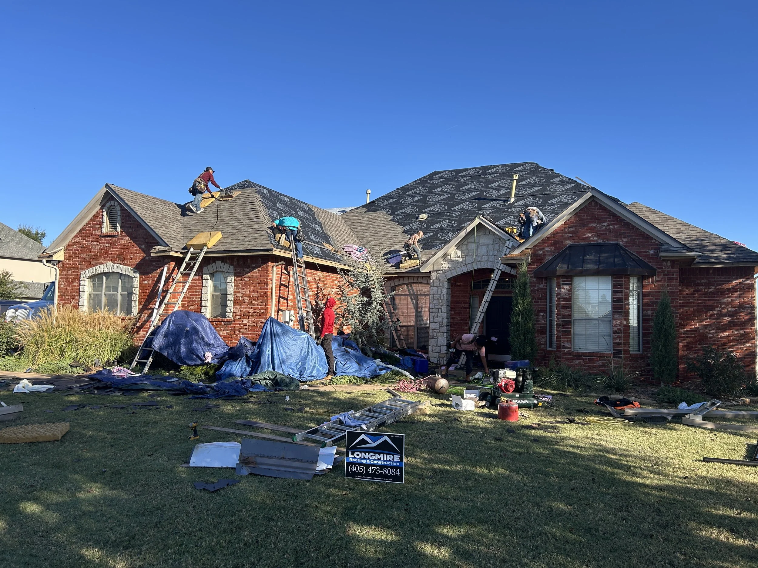 Construction workers on the roof of a brick house working on roof installation or repair, with equipment and tools scattered on the lawn below under a clear blue sky.