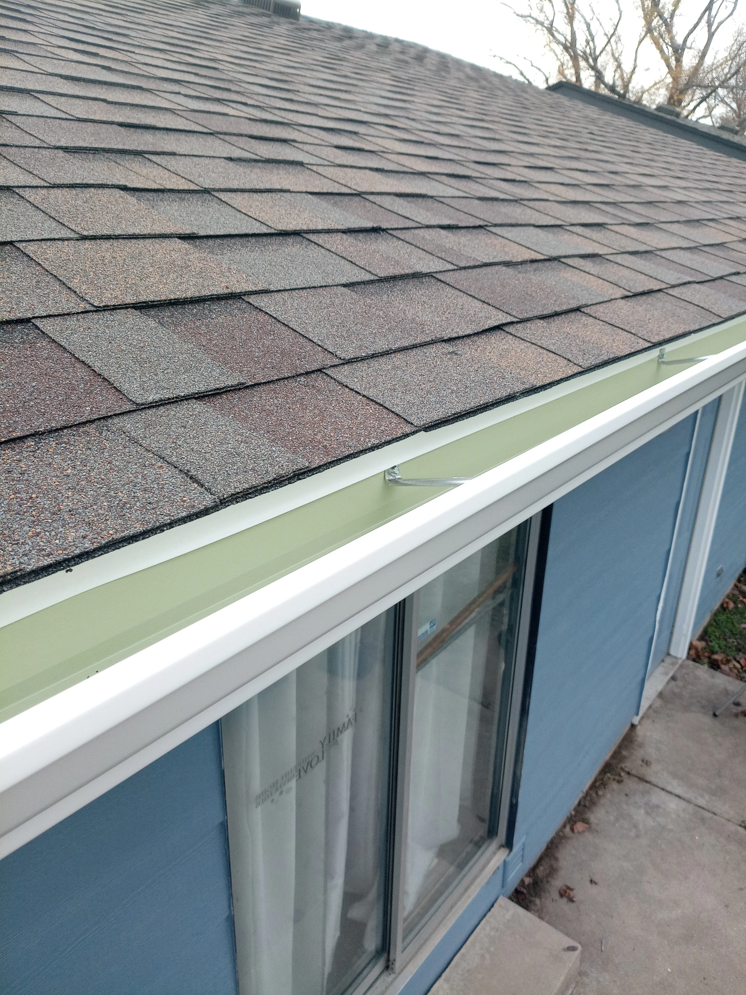Close up of a house's new gutters, new roof with brown shingles and a blue exterior wall with large windows, outside during daylight.