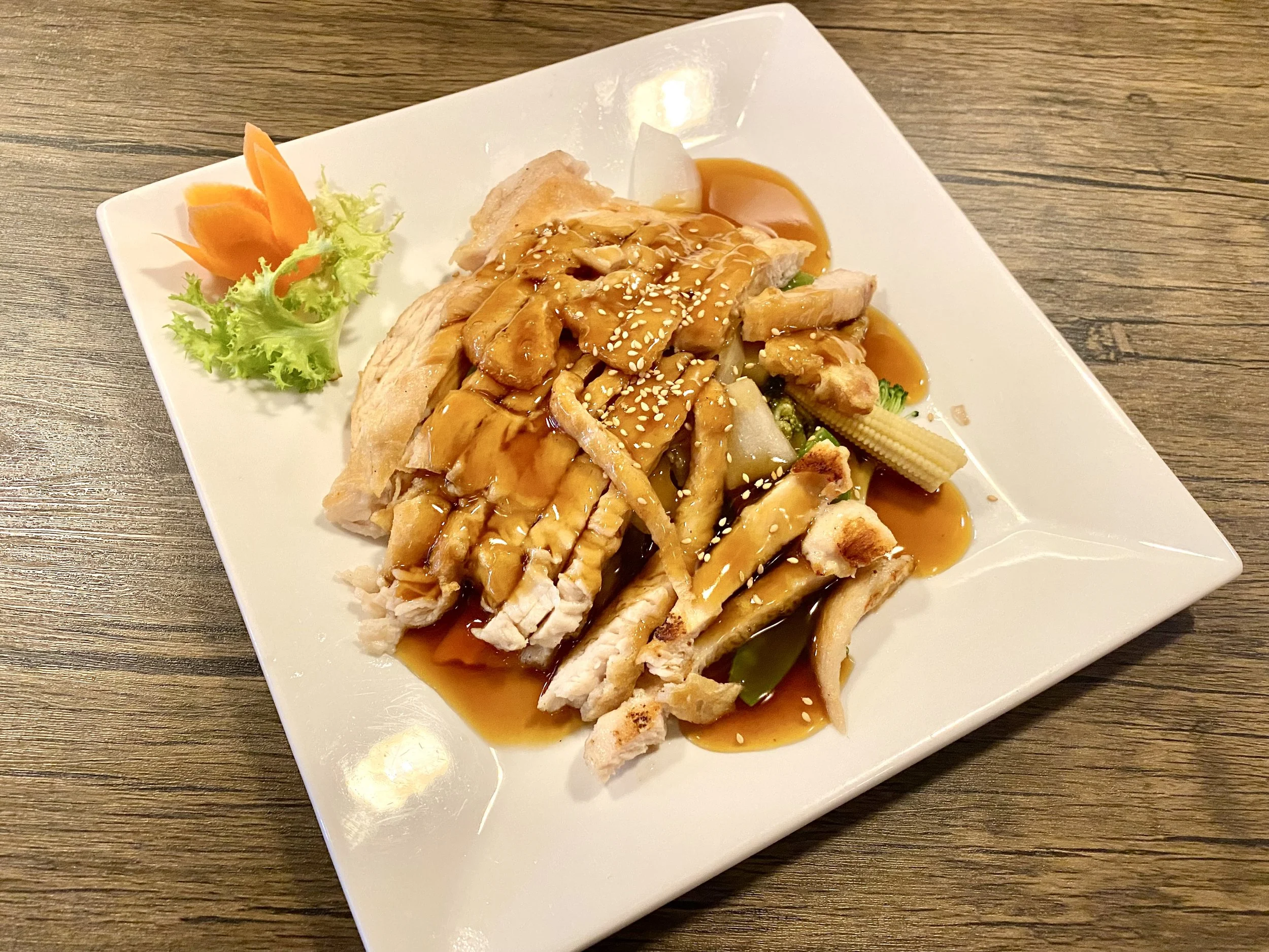 Plate with sliced grilled chicken, drizzled with brown sauce, garnished with sesame seeds, baby corn, lettuce, and a carved carrot flower on the side.
