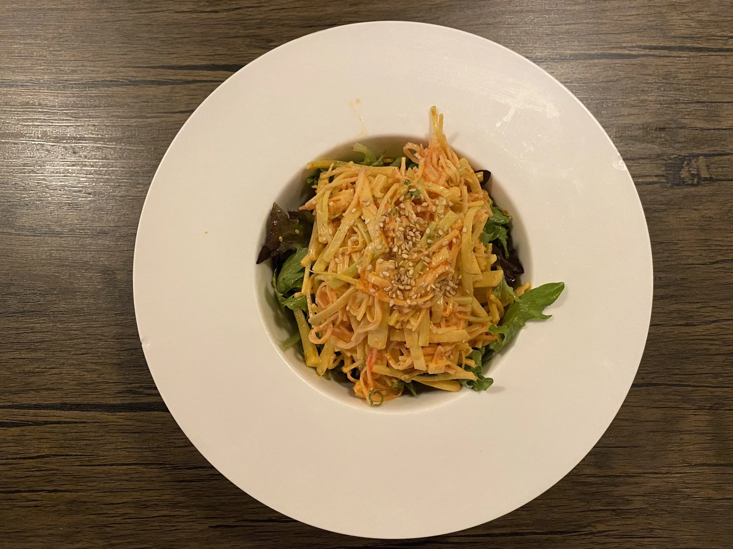 A white plate with a serving of mixed vegetable salad, topped with shredded carrots, sesame seeds, and julienned vegetables on a wooden table.