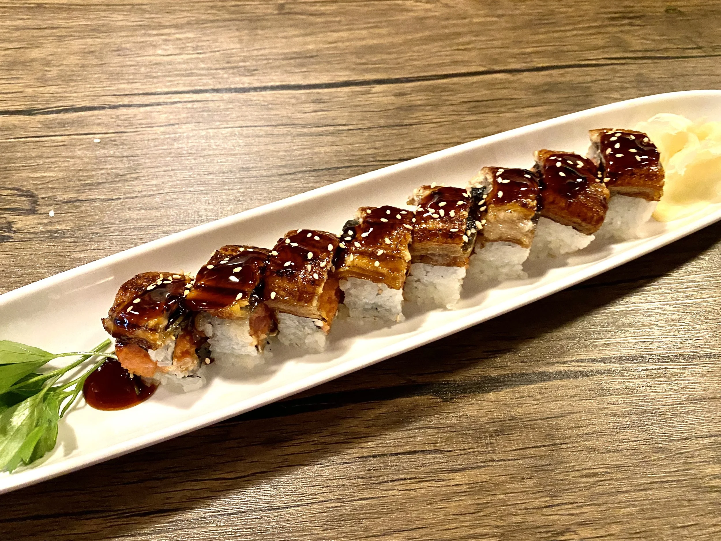 Sushi roll topped with eel sauce and sesame seeds, served with pickled ginger and wasabi on a long white plate.