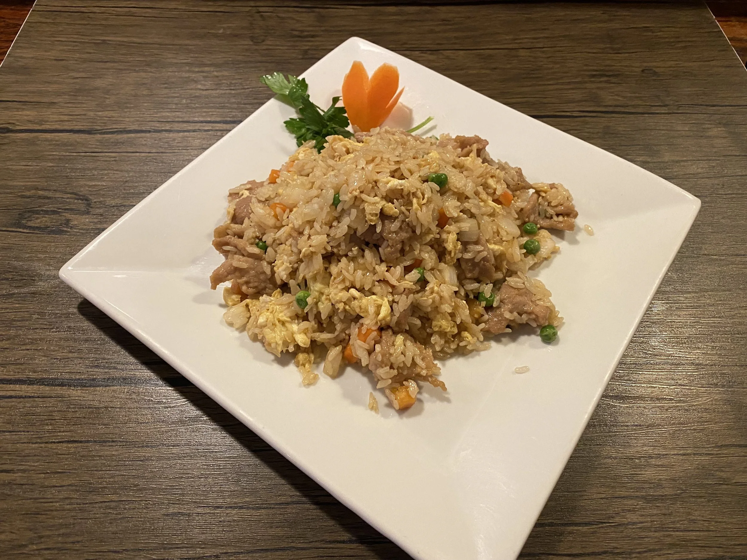 Plate of fried rice with peas, scrambled eggs, and bits of meat, garnished with a sprig of parsley and an orange carved flower on a white square plate.