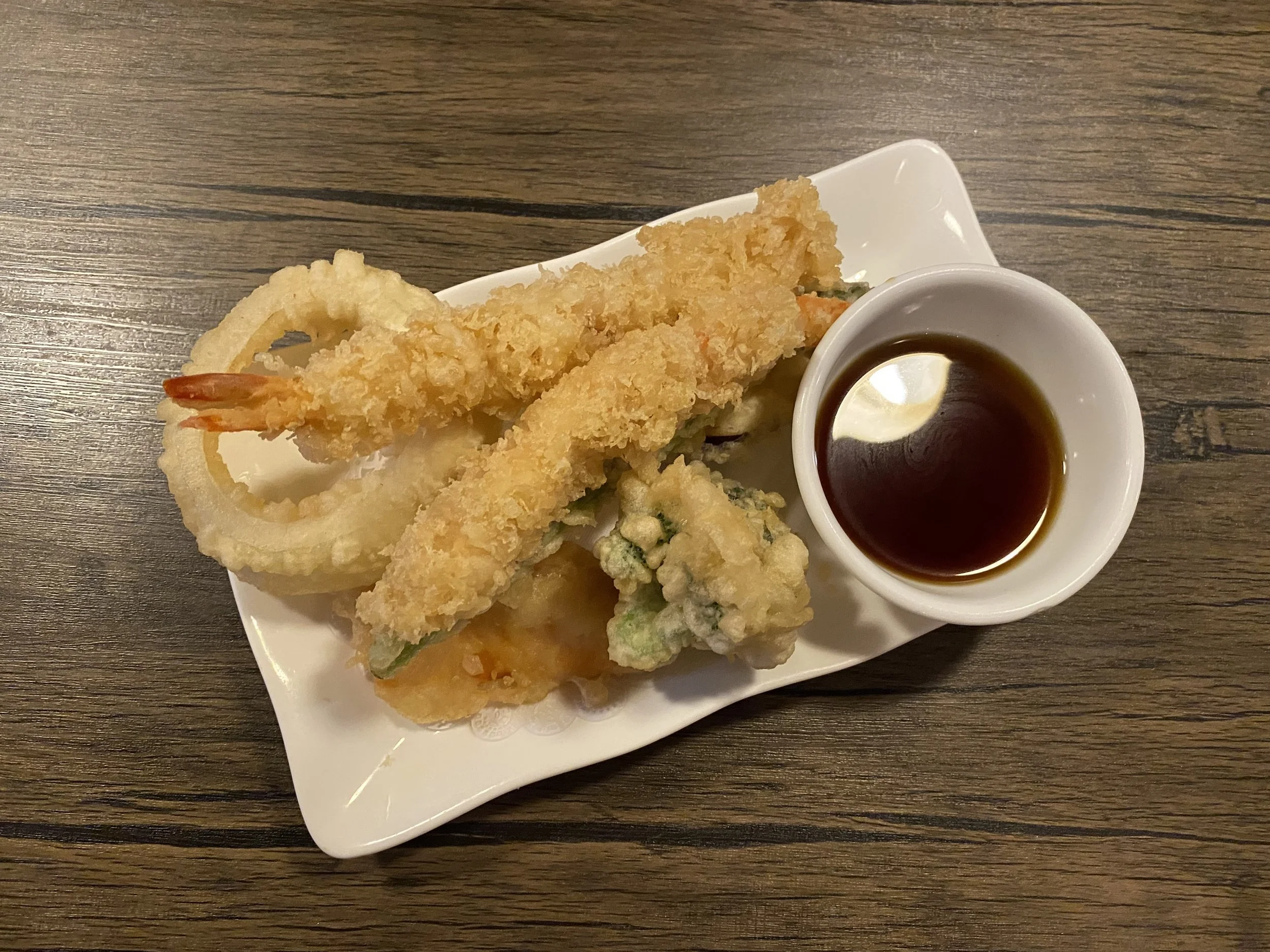 Assorted tempura on a white plate with a small bowl of dipping sauce, placed on a wooden table.