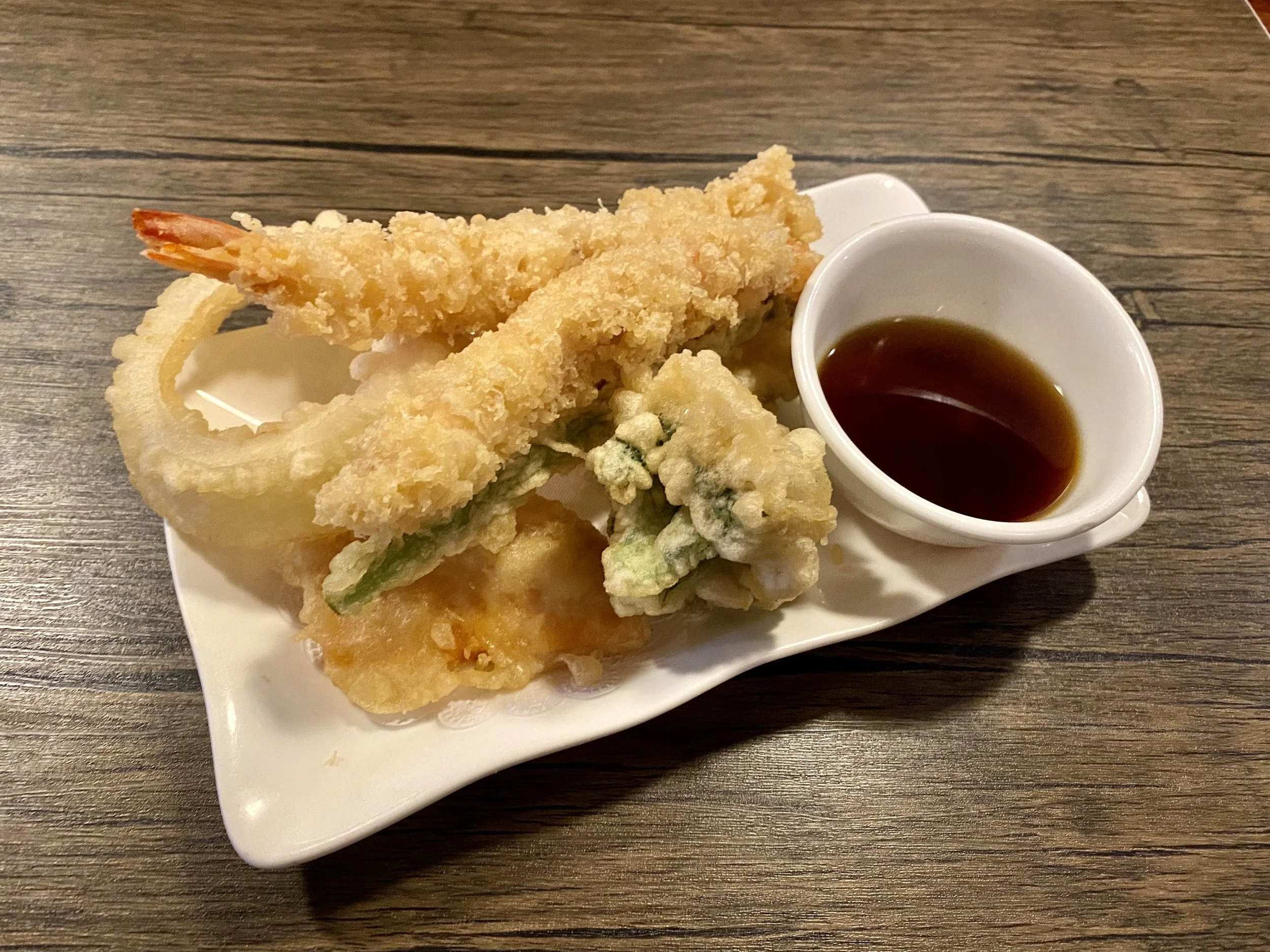 Assorted tempura with dipping sauce on a white plate placed on a wooden table.