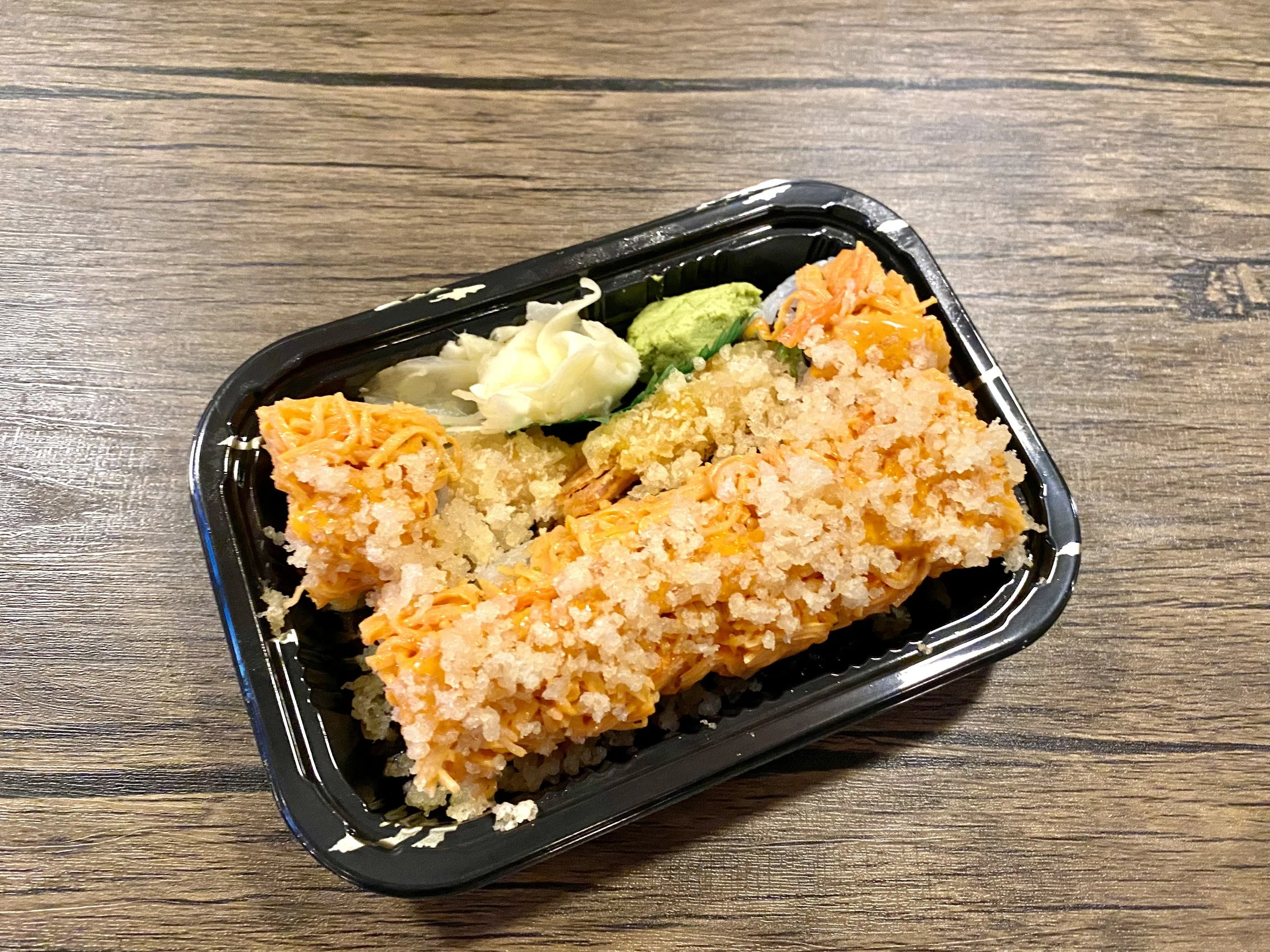 A black plastic takeout container with baked salmon topped with shredded cheese, served on a wooden table with garnish of shredded cabbage, lettuce, and guacamole.
