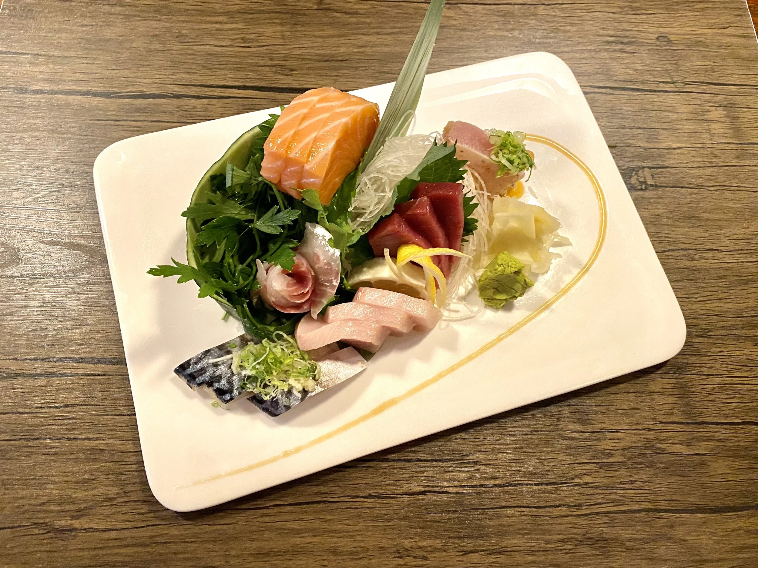 An assortment of sashimi including salmon, tuna, and mackerel, garnished with herbs and accompanied by wasabi, shredded daikon radish, and green onions, served on a white rectangular plate.