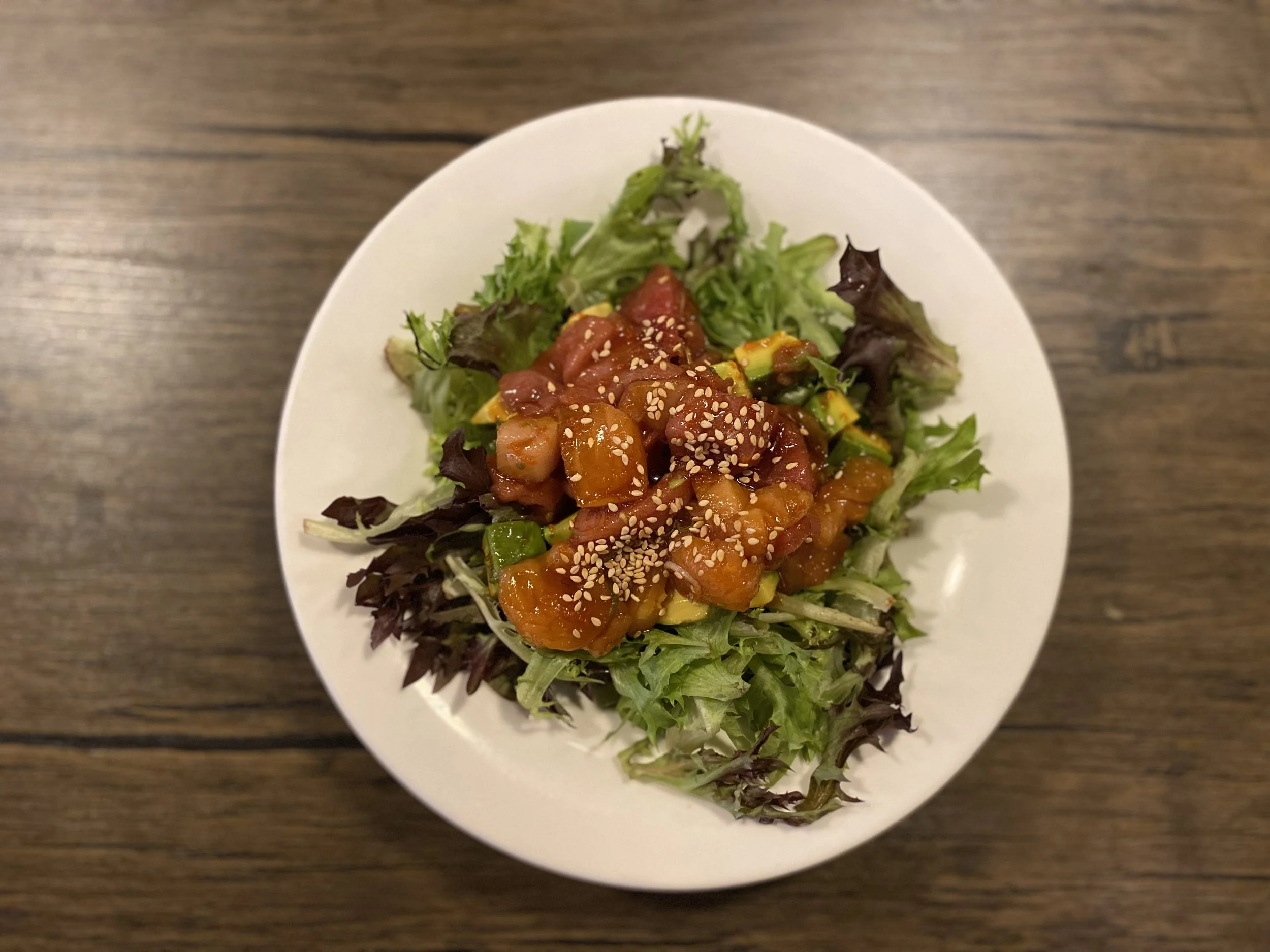 Mixed greens salad topped with chopped vegetables and sesame seeds, served on a white plate on a wooden table.