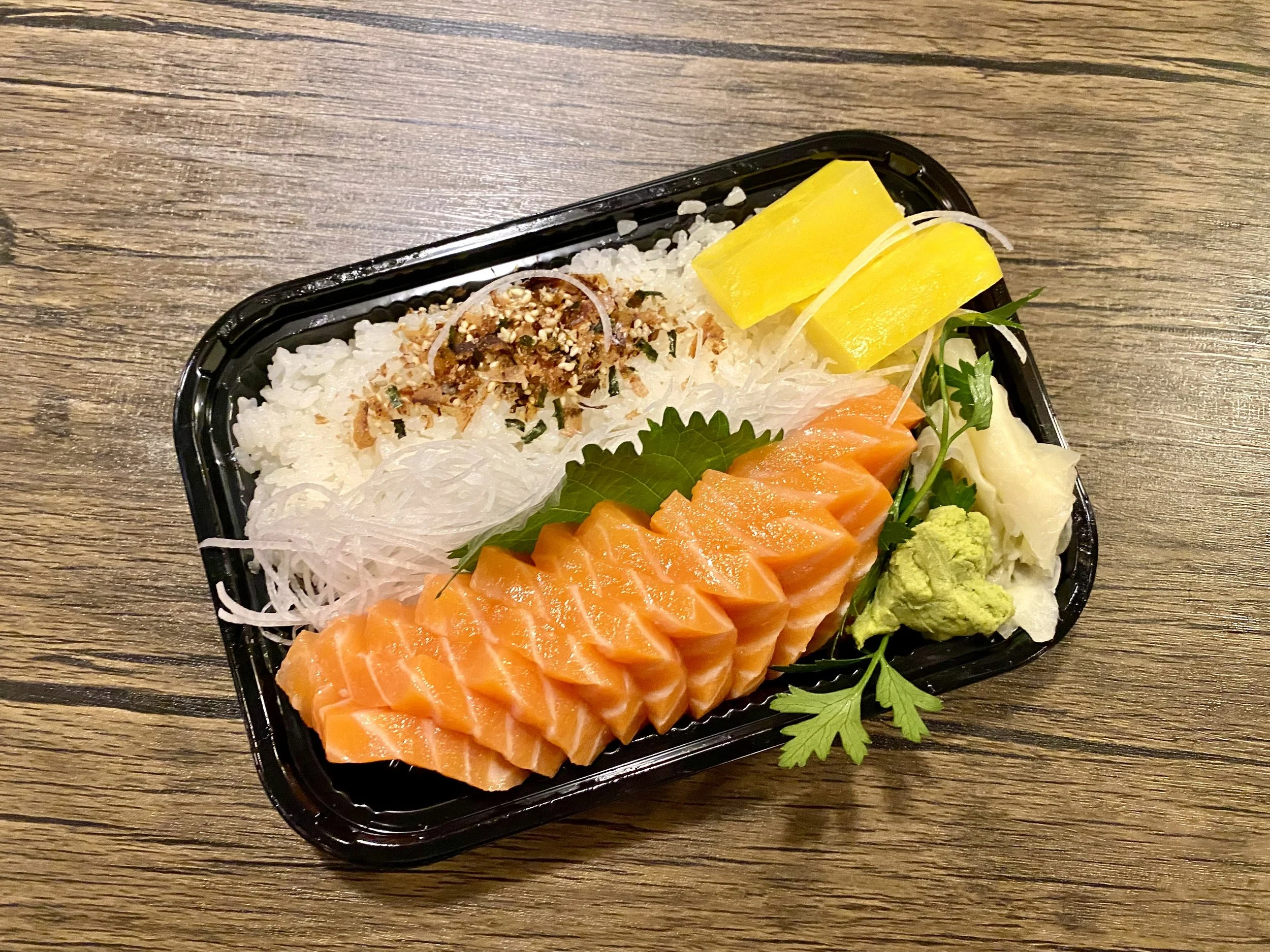A bento box with slices of salmon sashimi, white shredded daikon radish, yellow pickled radish, wasabi, and garnishes on a wooden table.