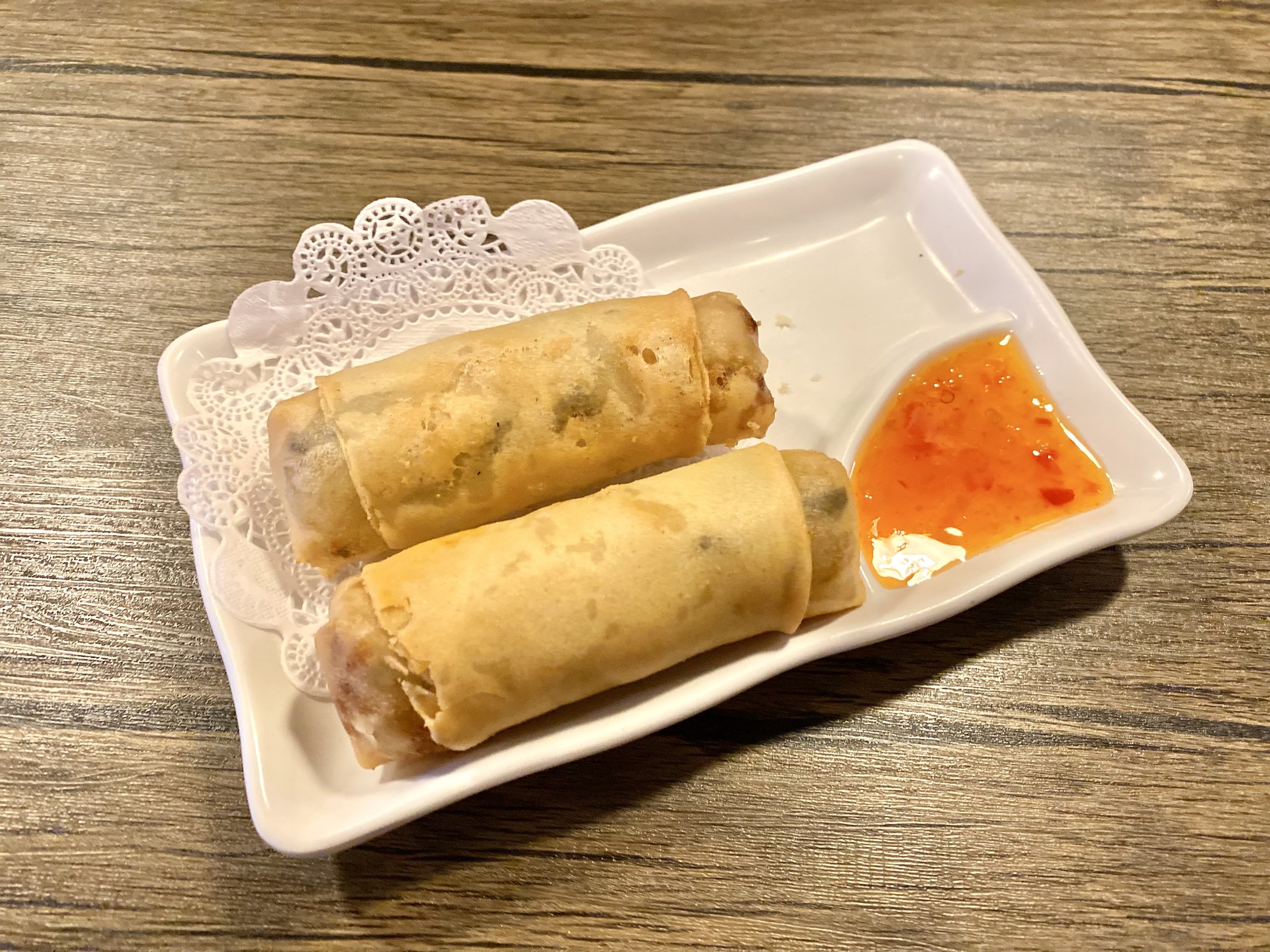 Two spring rolls on a white ceramic dish with a side of orange dipping sauce, placed on a wooden table.