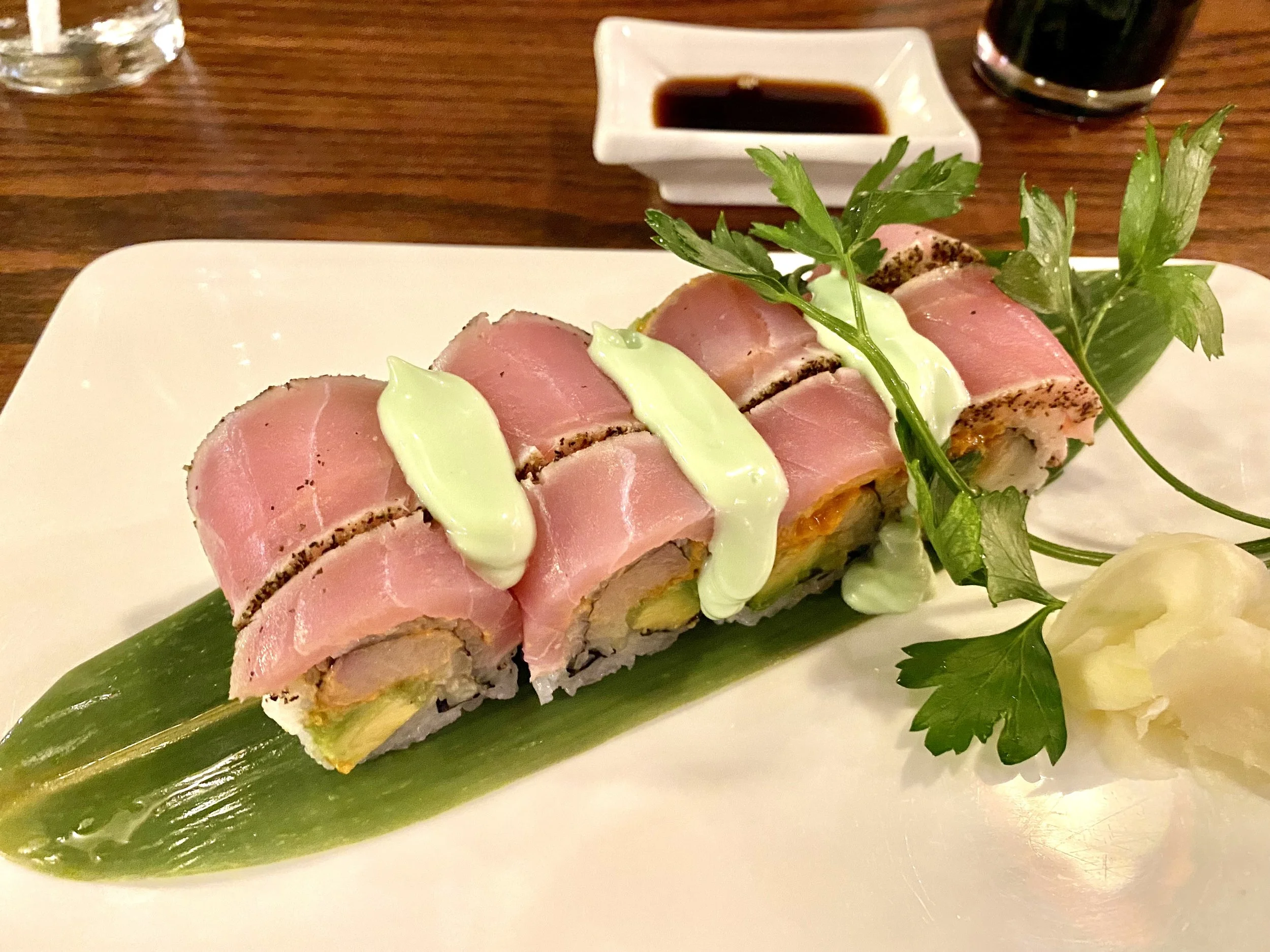 Sushi roll topped with slices of raw tuna, drizzled with a light green sauce, garnished with parsley, served on a leaf on a white plate, with pickled ginger, soy sauce in a small dish, and a dark drink in the background.