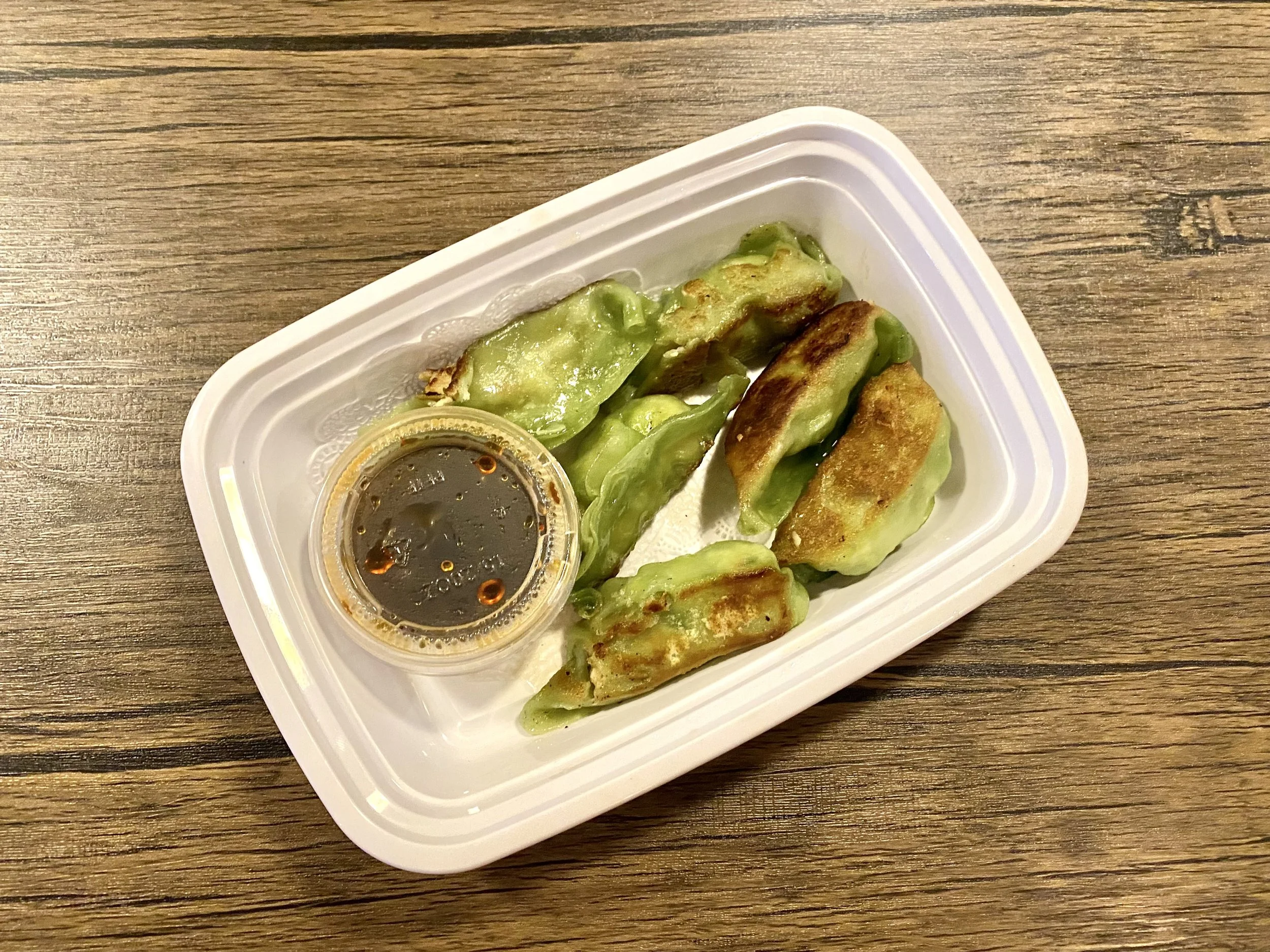 Six cooked dumplings with soy sauce in a small container, served on a white plastic tray on a wooden table.