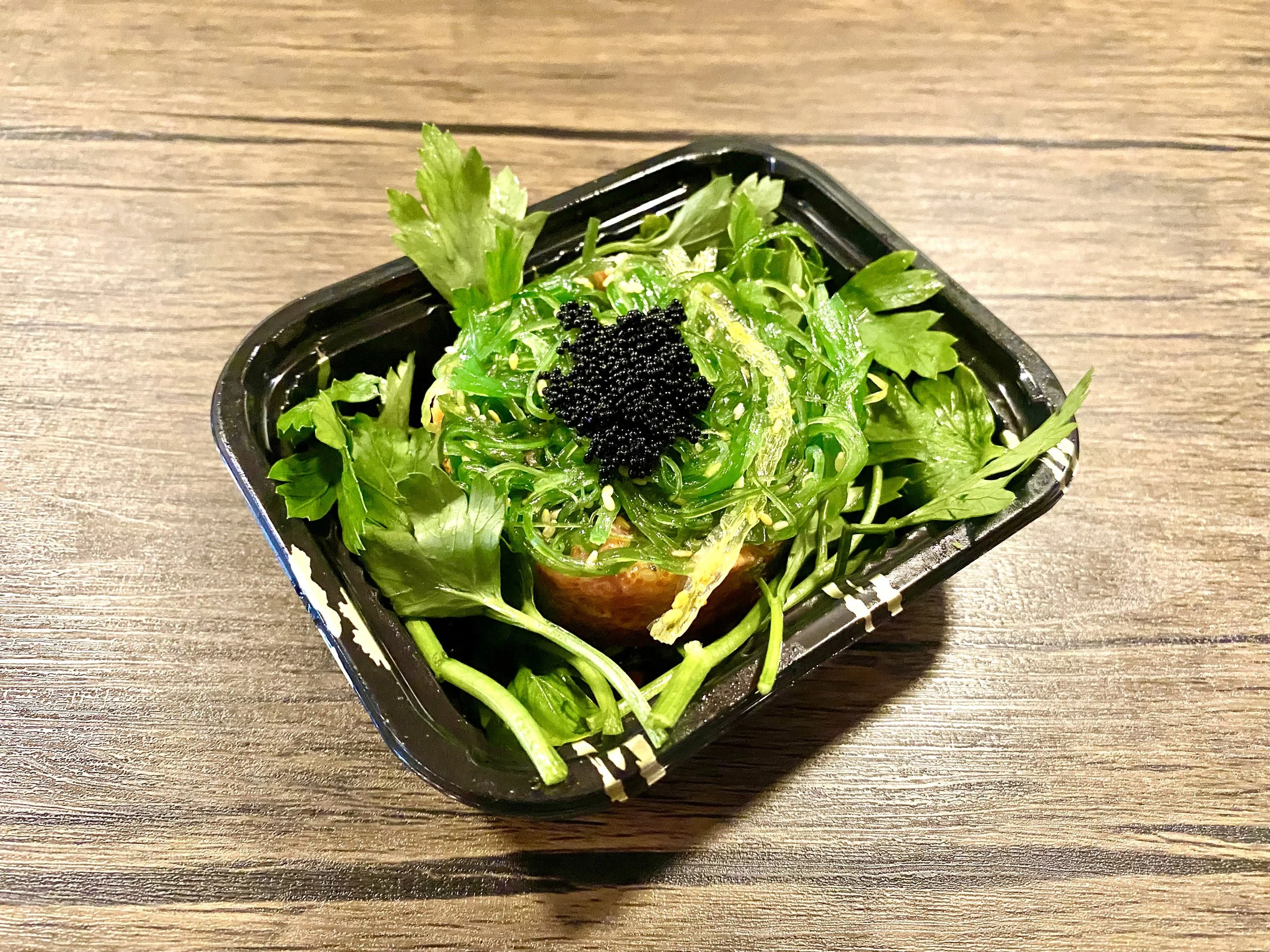 A small black container of salad with seaweed and green herbs on a wooden surface.