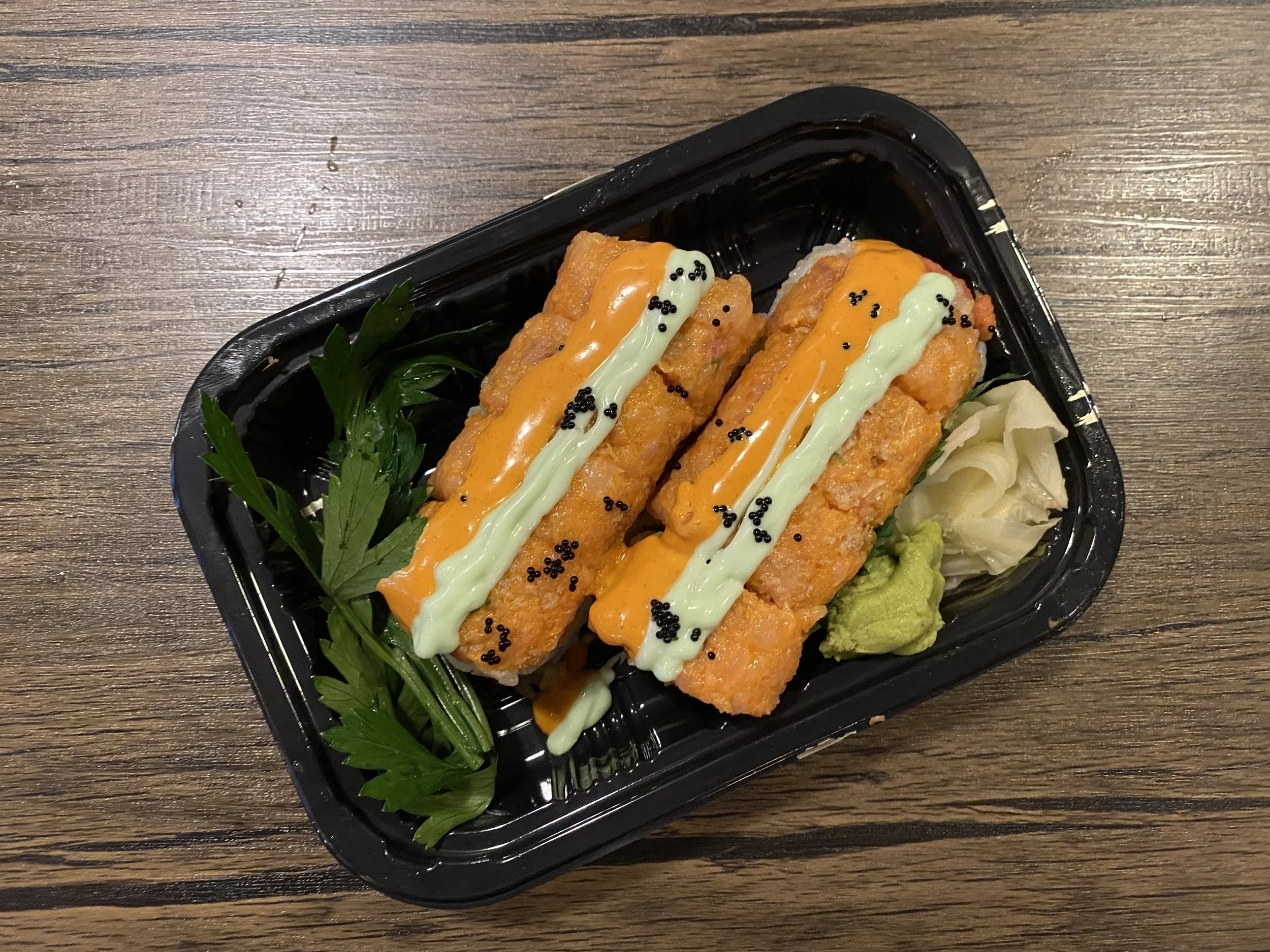 Two pieces of sushi topped with orange sauce and green sauce, garnished with black sesame seeds, served with wasabi, pickled ginger, and green leaf on a black tray.