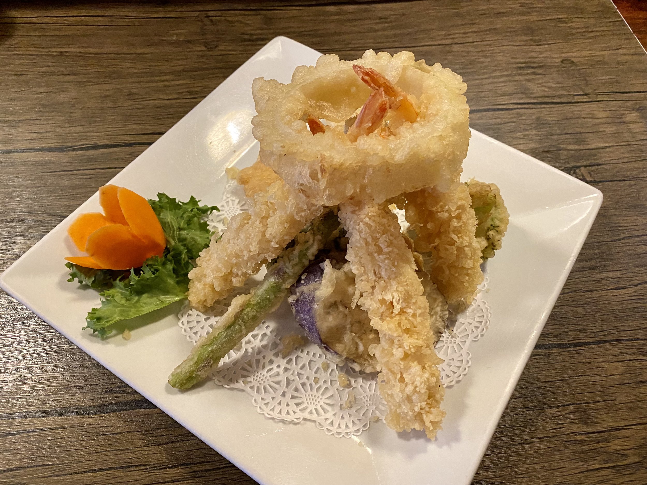 Assorted tempura fried vegetables and shrimp on a white plate garnished with lettuce and an edible flower, placed on a wooden table.