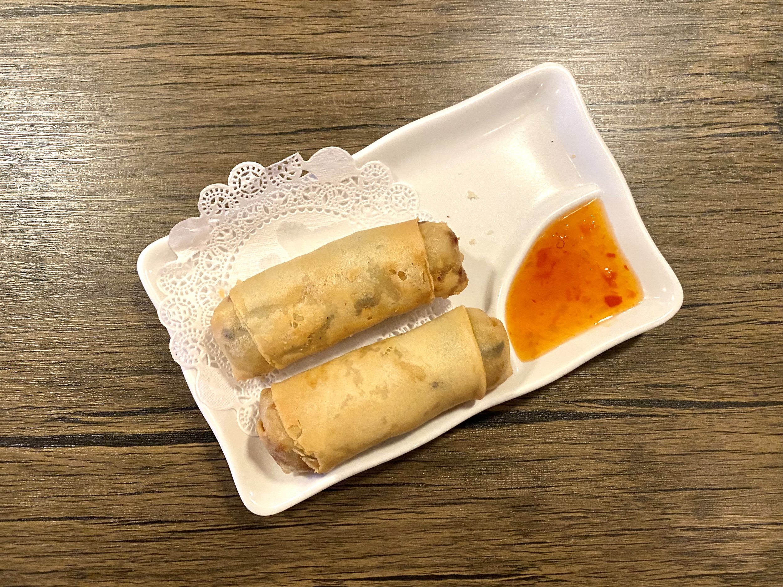 Two spring rolls on a white plate with a small side of chili sauce, placed on a wooden table.