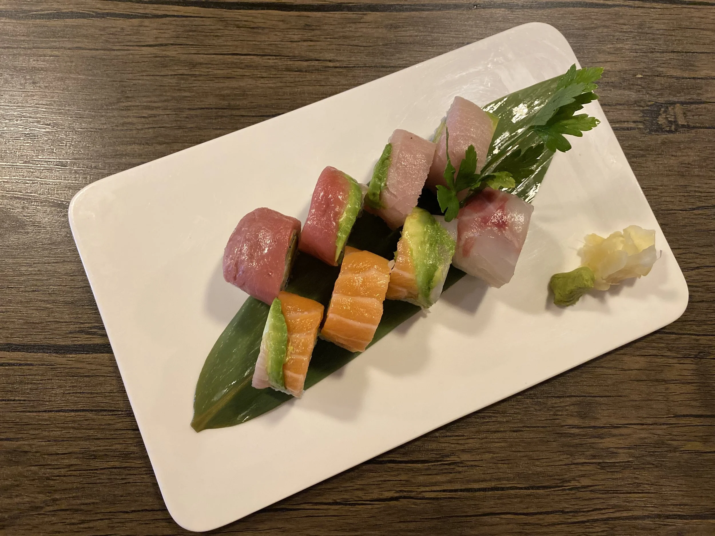 Assorted slices of raw fish and seafood on a white rectangular plate with a green leaf, wasabi, and ginger, garnished with a sprig of parsley, served on a wooden table.